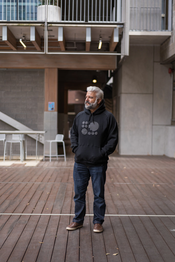 Full view of the Puff Pixel Hoodie in black, worn by a model standing on RMIT campus with hands in pockets. The hoodie features raised grey pixel patterns across the chest. The model is wearing blue jeans. Available at RMIT Store.