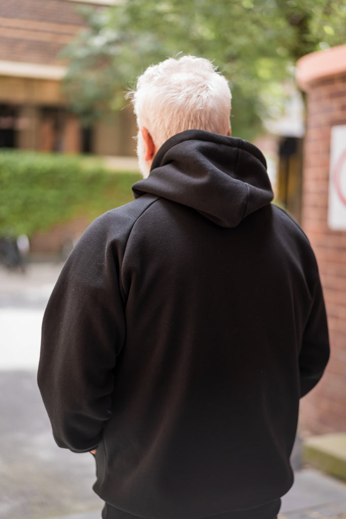 Rear view of the Puff Pixel Hoodie in black, showing a plain back without any logos or patterns. Model is standing outdoors on campus, with the hoodie’s relaxed fit and hood shape clearly visible. Available at RMIT Store.