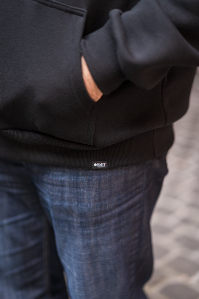 Zoomed-in view of the bottom edge of the Puff Pixel Hoodie, showing the raised white RMIT pixel logo printed on the black fabric. The model is wearing blue jeans, creating a clear contrast with the hoodie. Available at RMIT Store.