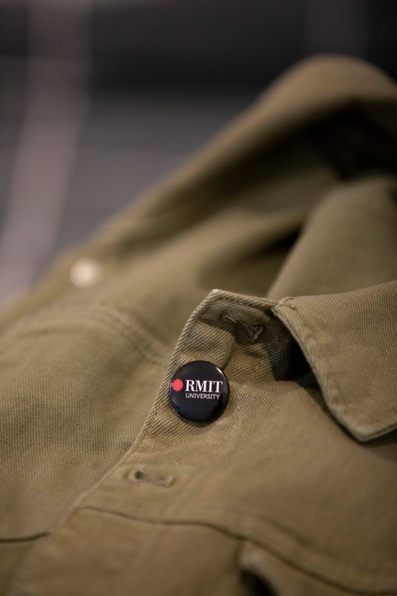 A black RMIT University badge on a green jacket. The badge features the RMIT logo in white and red, clearly visible on the left chest area of the jacket.