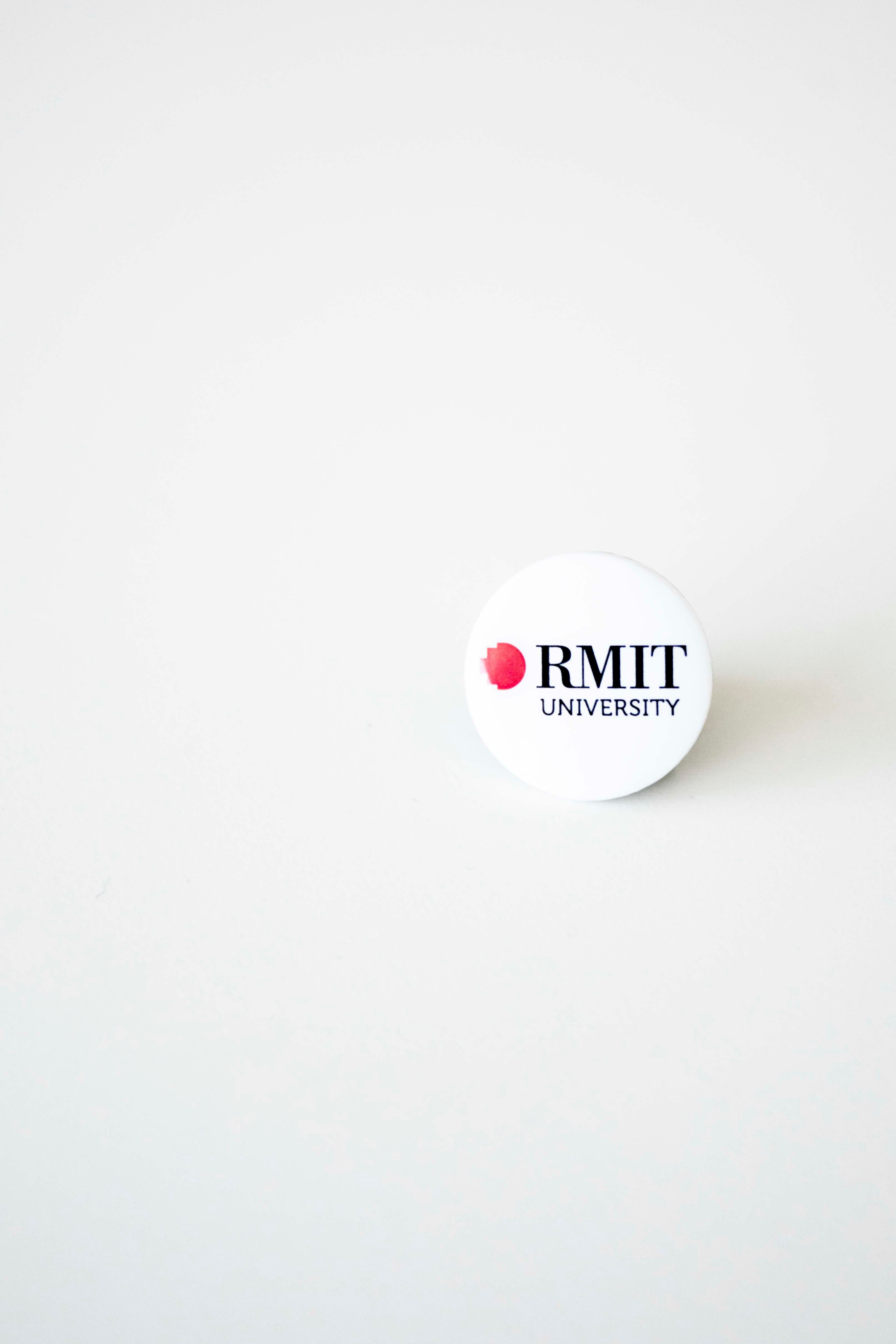 Front view of an RMIT University badge in white. The badge prominently displays the RMIT logo in bold, clear text, suitable for attaching to clothing or accessories.