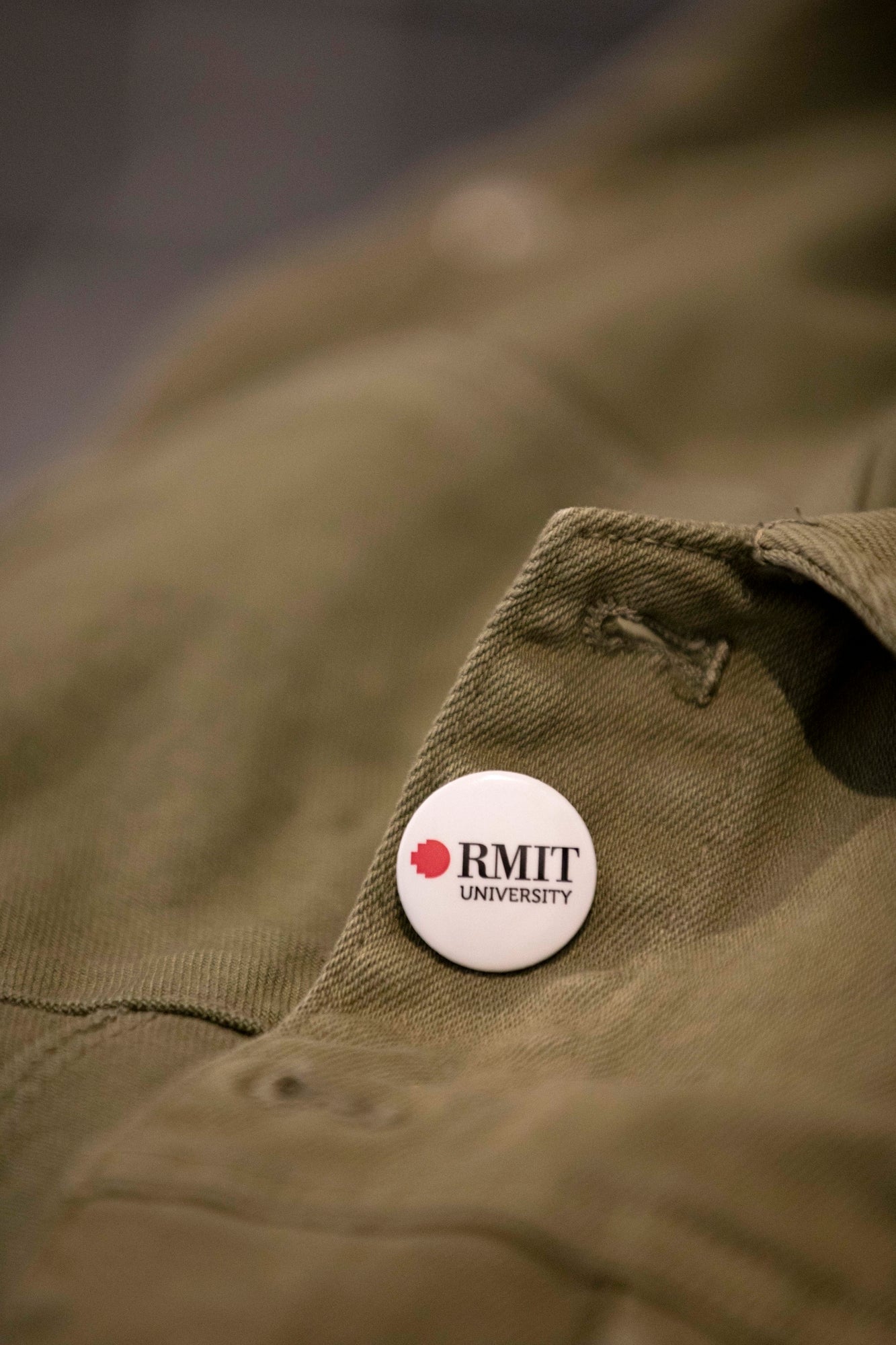 A white RMIT University badge on a green jacket. The badge displays the RMIT logo in white and red, prominently positioned on the left chest area of the jacket.