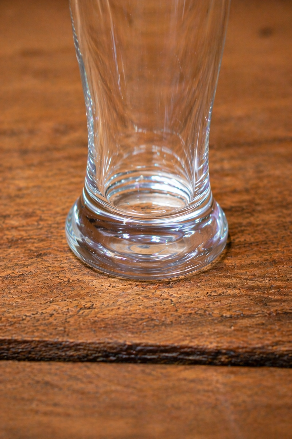 Close-up detail of the RMIT Beer Pint Gift Pack showing the thick, weighted clear glass base with smooth curved edges and subtle concentric mould lines, positioned on a warm light grey wooden tabletop surface, highlighting the solid construction and clarity of the glass available at the RMIT Store.