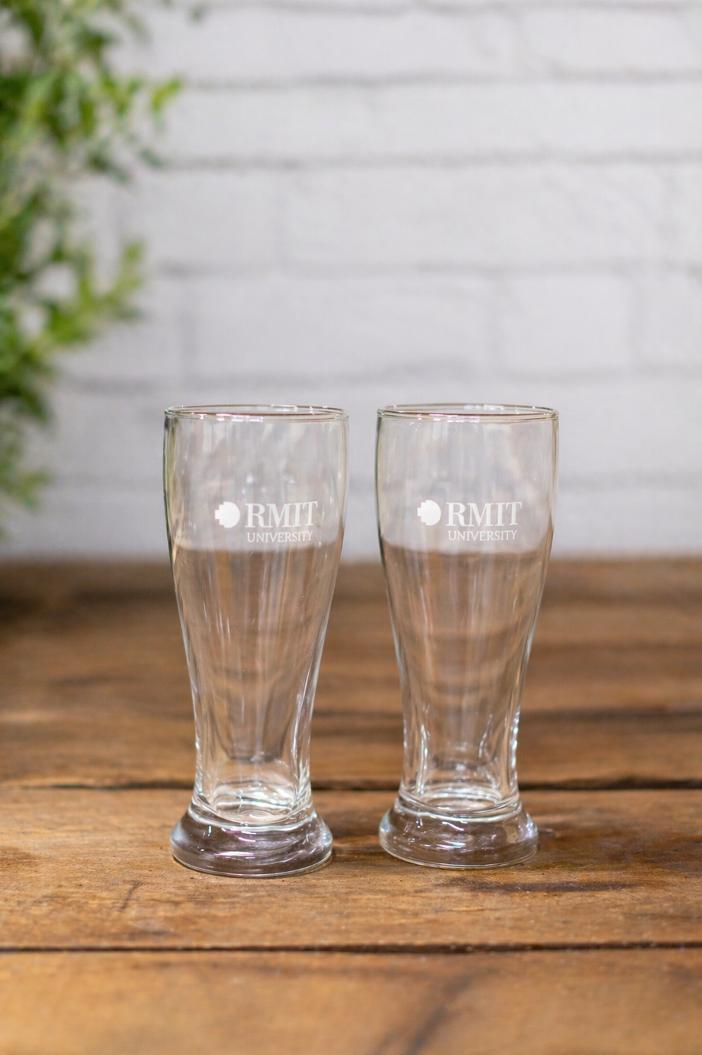 RMIT Beer Pint Gift Pack showing two clear glass beer glasses with gently curved sides and sturdy bases, each printed with a white RMIT University logo on the front, positioned evenly on a light grey wooden tabletop against a soft neutral background, emphasising the matching pair design available at the RMIT Store.