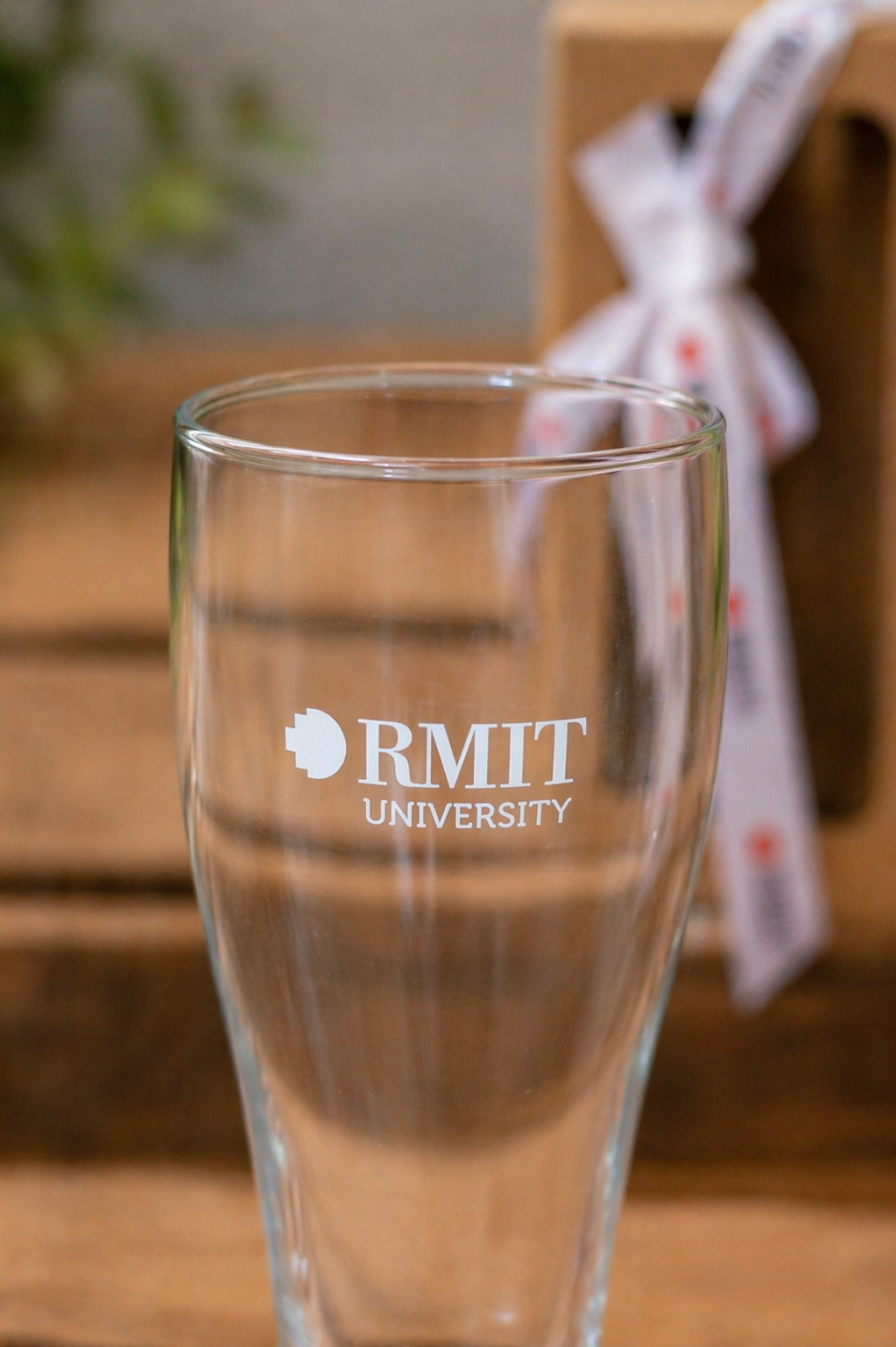 RMIT Beer Pint Gift Pack clear tapered beer glass featuring a crisp white printed RMIT University logo with circular emblem and text centred on the front, shown upright on a light grey wooden tabletop with a blurred gift box and ribbon in the background, clearly displaying branding placement available at the RMIT Store.