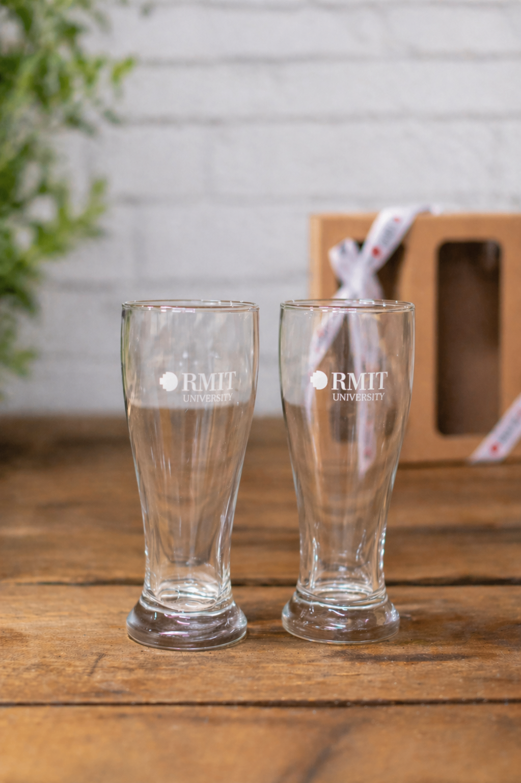 RMIT Beer Pint Gift Pack featuring two identical clear tapered beer glasses with thick bases and white RMIT University logos facing forward, displayed side by side on a light grey wooden tabletop with a kraft gift box and branded ribbon softly blurred in the background, highlighting the two-piece set available at the RMIT Store.