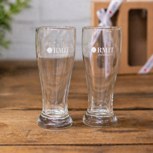 RMIT Beer Pint Gift Pack featuring two identical clear tapered beer glasses with thick bases and white RMIT University logos facing forward, displayed side by side on a light grey wooden tabletop with a kraft gift box and branded ribbon softly blurred in the background, highlighting the two-piece set available at the RMIT Store.
