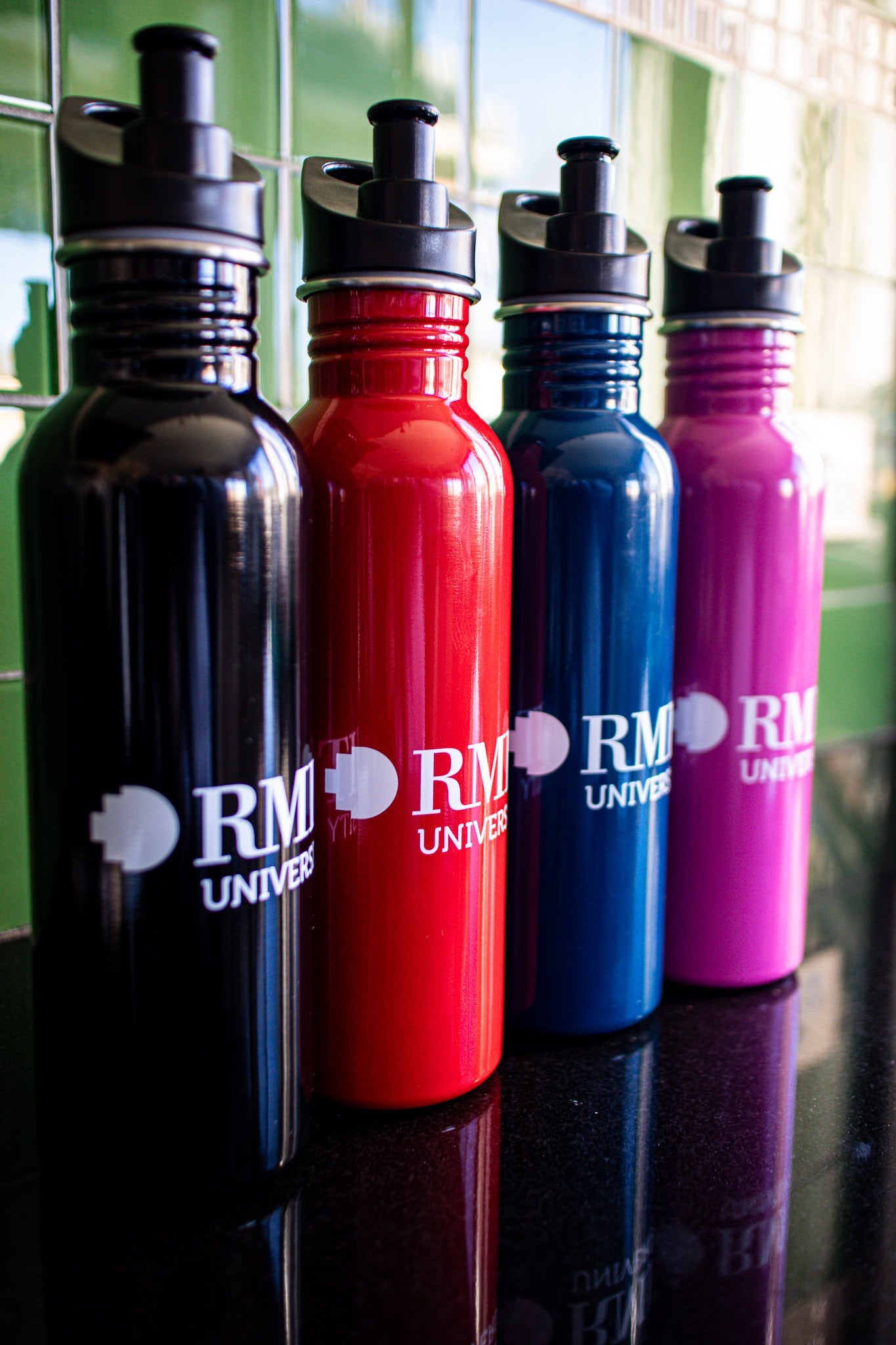 A collection of four RMIT bottles in red, black, navy, and pink, arranged in an angled line on a black tabletop with a green background. Each bottle prominently features the white RMIT University logo on its side. This image showcases the colour variety and branding, available at the RMIT Store.