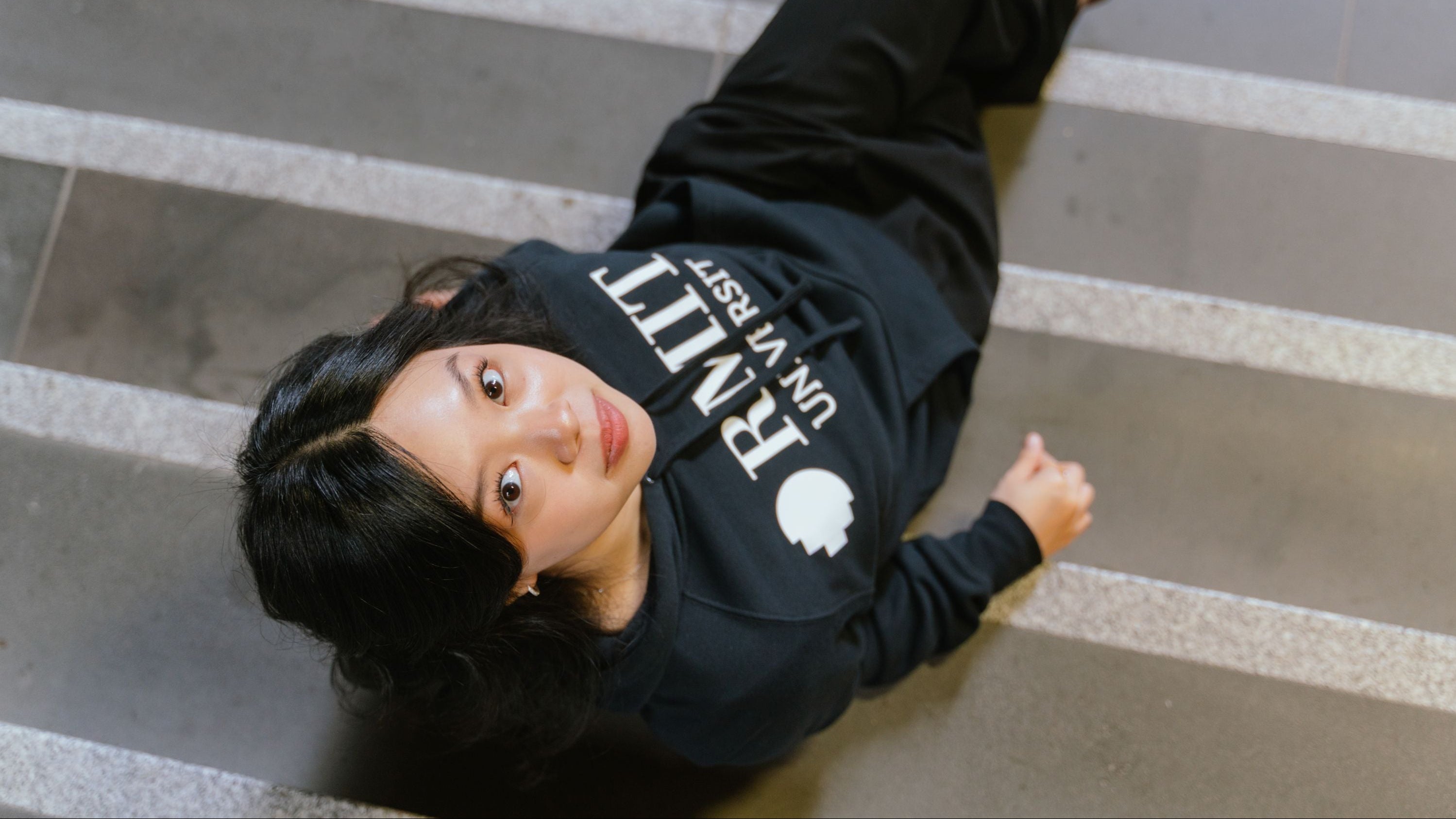 Top-down view of model lying on indoor staircase wearing RMIT Essential Hoodie in navy featuring bold white RMIT University logo across the chest, with hood down and relaxed fit, captured from above with strong perspective lines of steps, available at the RMIT Store.