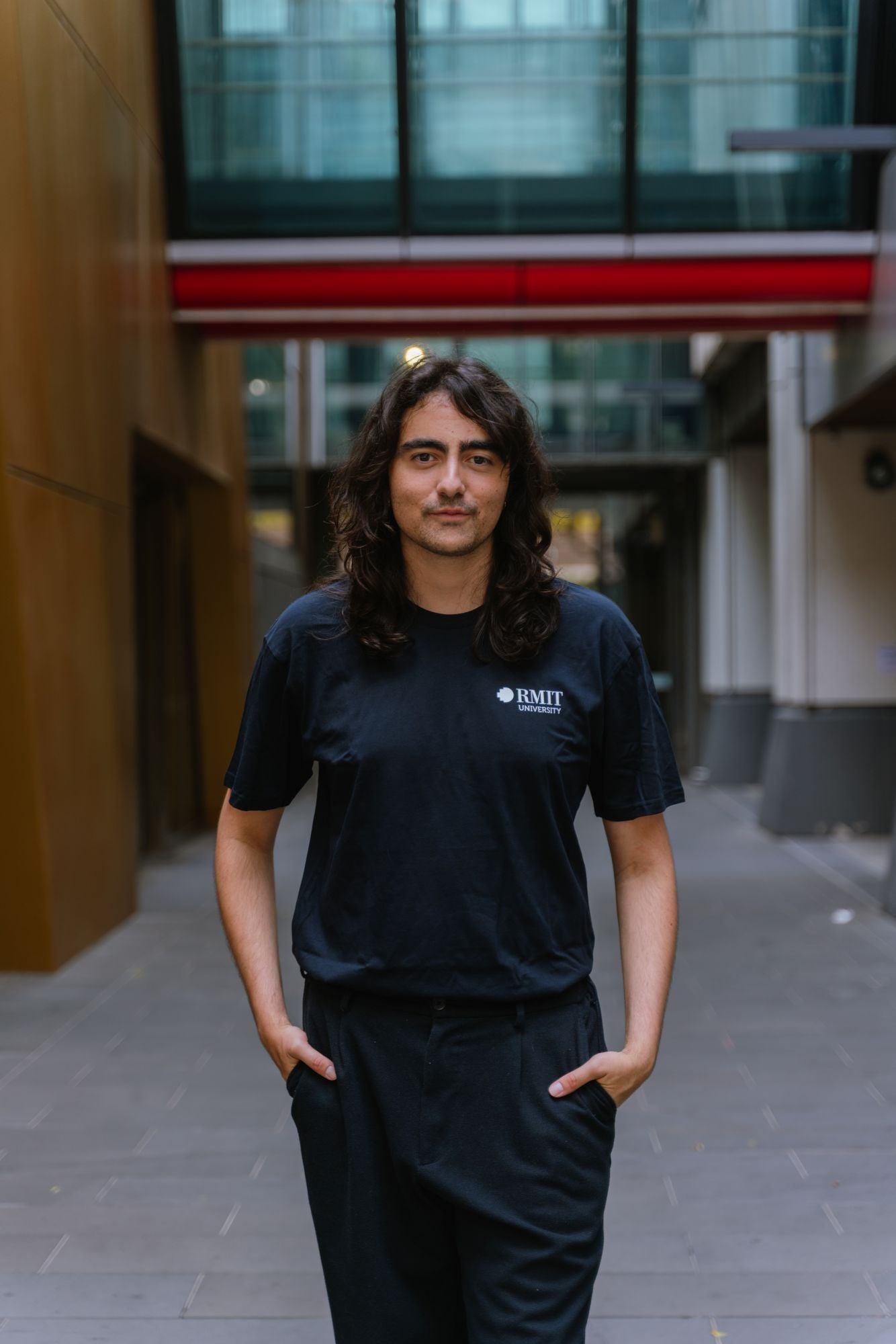 Model standing in an outdoor campus walkway wearing RMIT Essential Tee in navy featuring a small white RMIT University logo printed on the left chest, relaxed classic fit with short sleeves, paired with black pants and hands in pockets in a modern architectural setting, available at the RMIT Store.