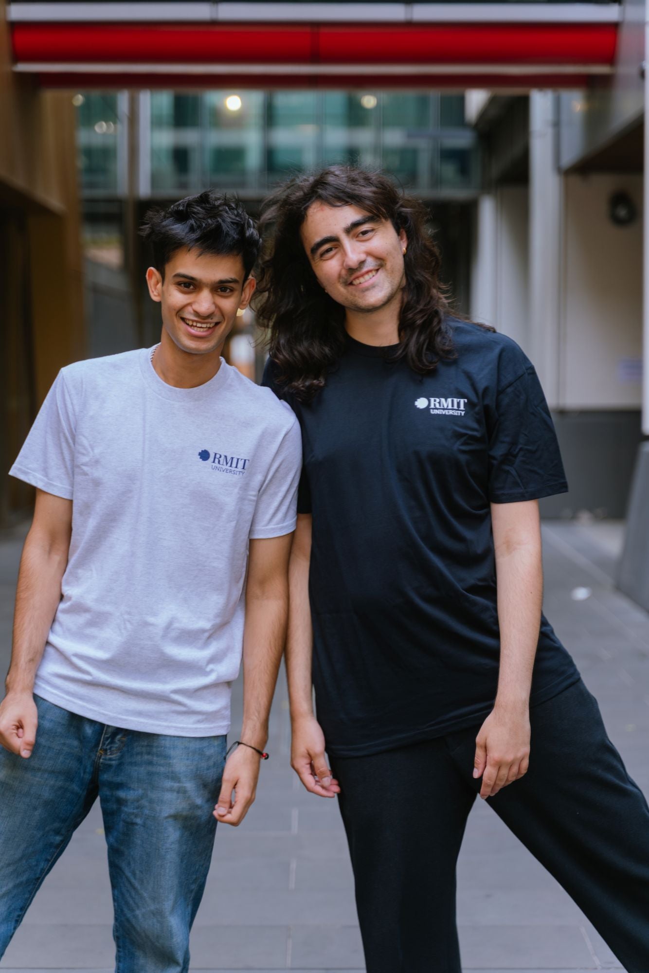 Two models standing outdoors in a modern campus walkway wearing RMIT Essential Tee, one in White Marle and one in Navy, both featuring a small RMIT University logo printed on the left chest; the model in White Marle tee wears blue denim jeans while the model in Navy tee wears black pants, both smiling and leaning slightly towards each other with blurred architectural background, available at the RMIT Store.