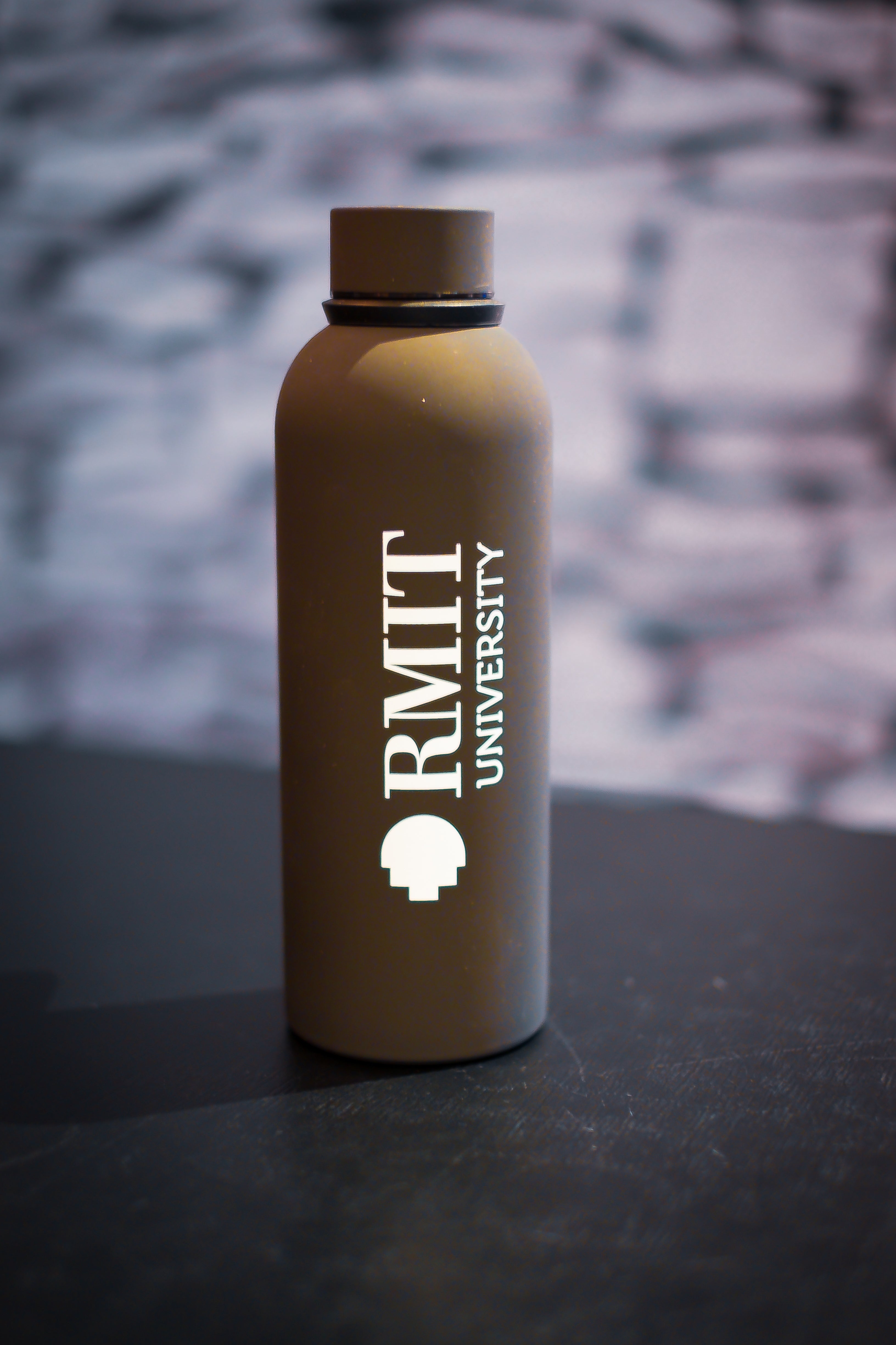 RMIT Everyday Bottle in matte black with screw-top lid, displayed upright on a black tabletop at RMIT Store, showing vertically printed white RMIT University logo and shield icon on the front, smooth powder-coated finish, cylindrical shape with rounded shoulders, and softly blurred stone-textured background.