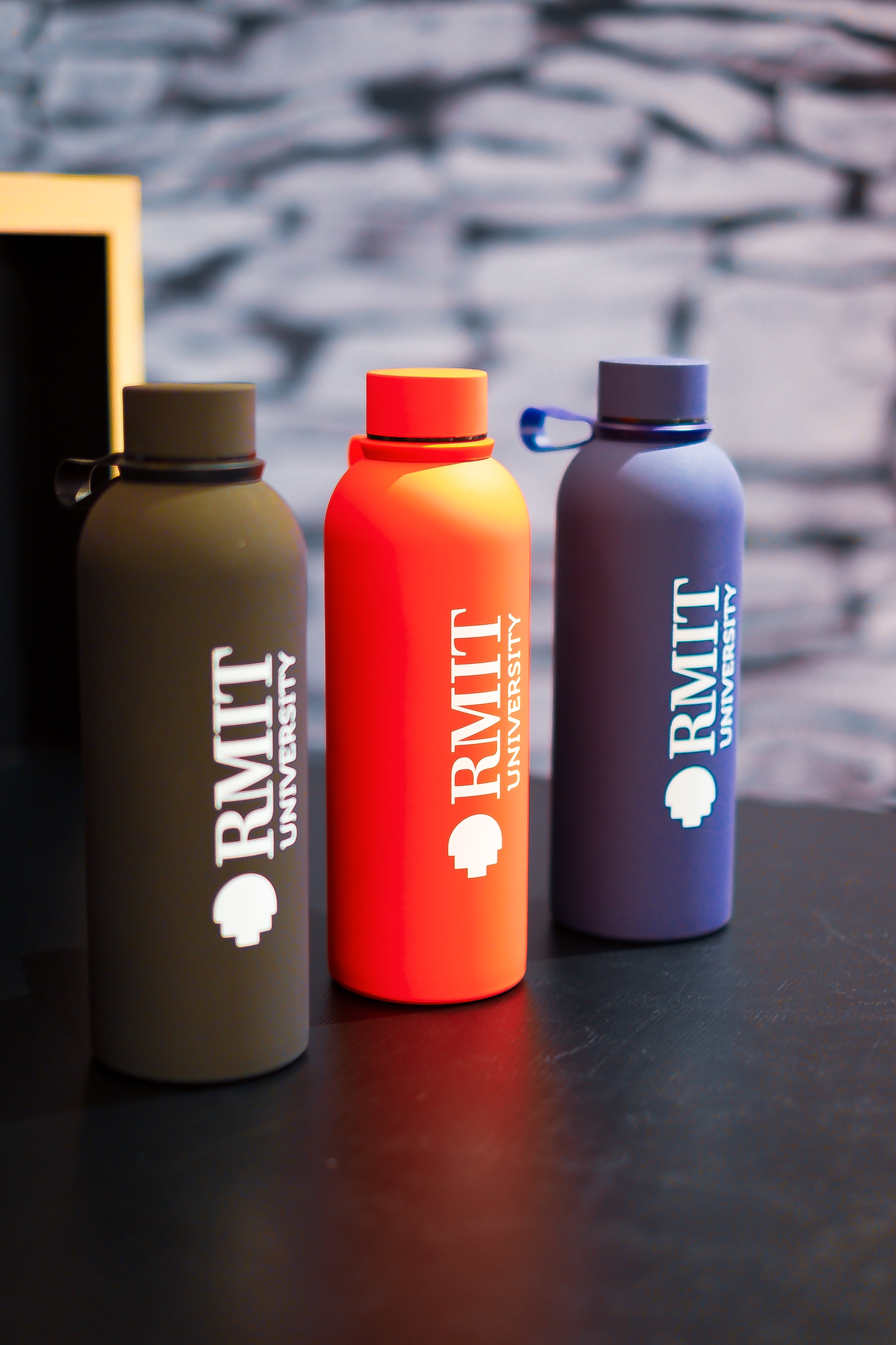 RMIT Everyday Bottle range displayed together at RMIT Store, showing three upright bottles in black, red, and navy blue with vertically printed white RMIT University logos, matching screw-top lids with carry loops, arranged on a black tabletop against a textured stone-style background.