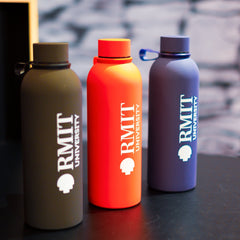 RMIT Everyday Bottle range displayed together at RMIT Store, showing three upright bottles in black, red, and navy blue with vertically printed white RMIT University logos, matching screw-top lids with carry loops, arranged on a black tabletop against a textured stone-style background.