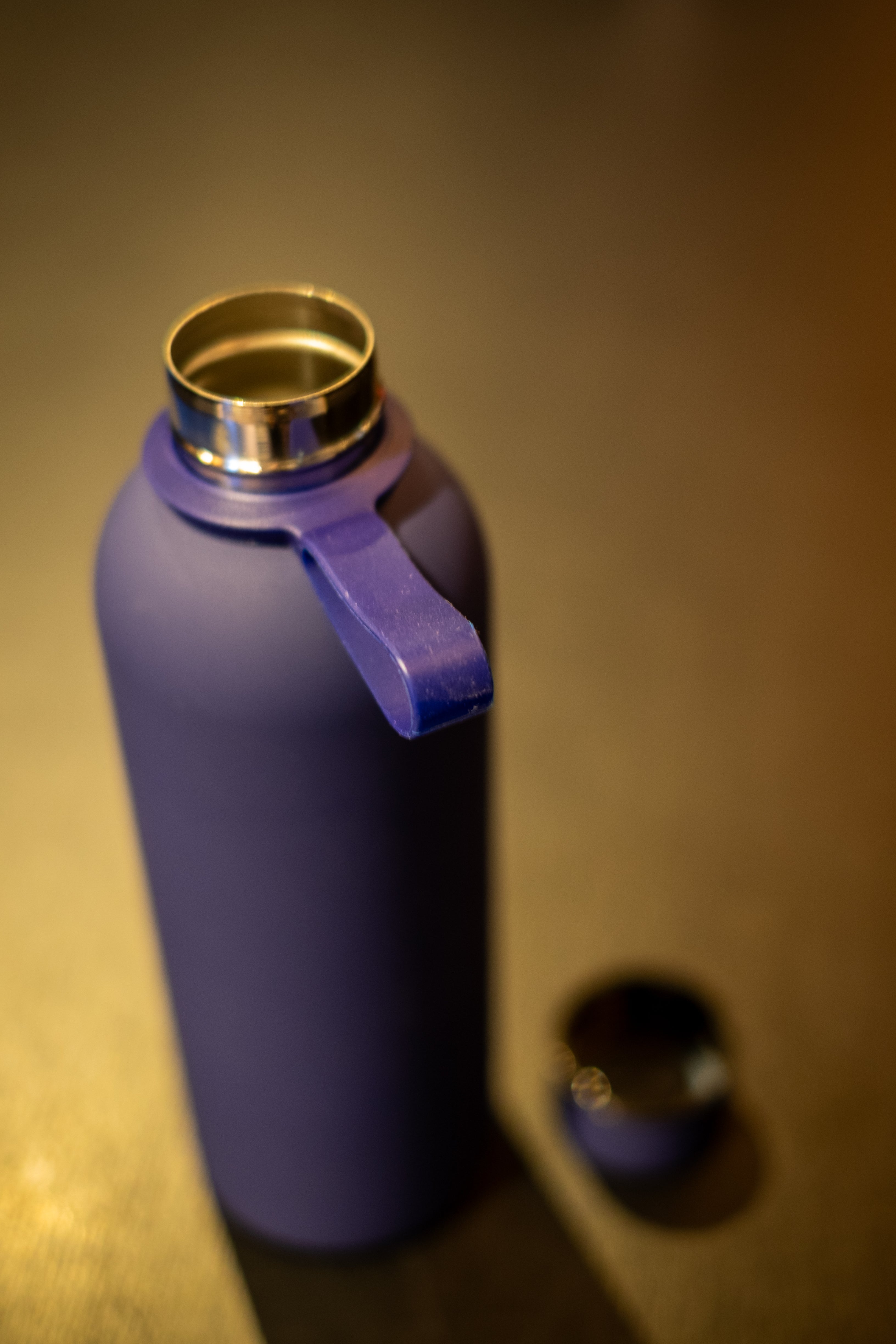 RMIT Everyday Bottle in matte navy blue with integrated silicone carry loop, shown open on a wooden surface at RMIT Store, revealing stainless steel threaded neck and reflective interior rim, with detached matching lid placed nearby and softly blurred background.