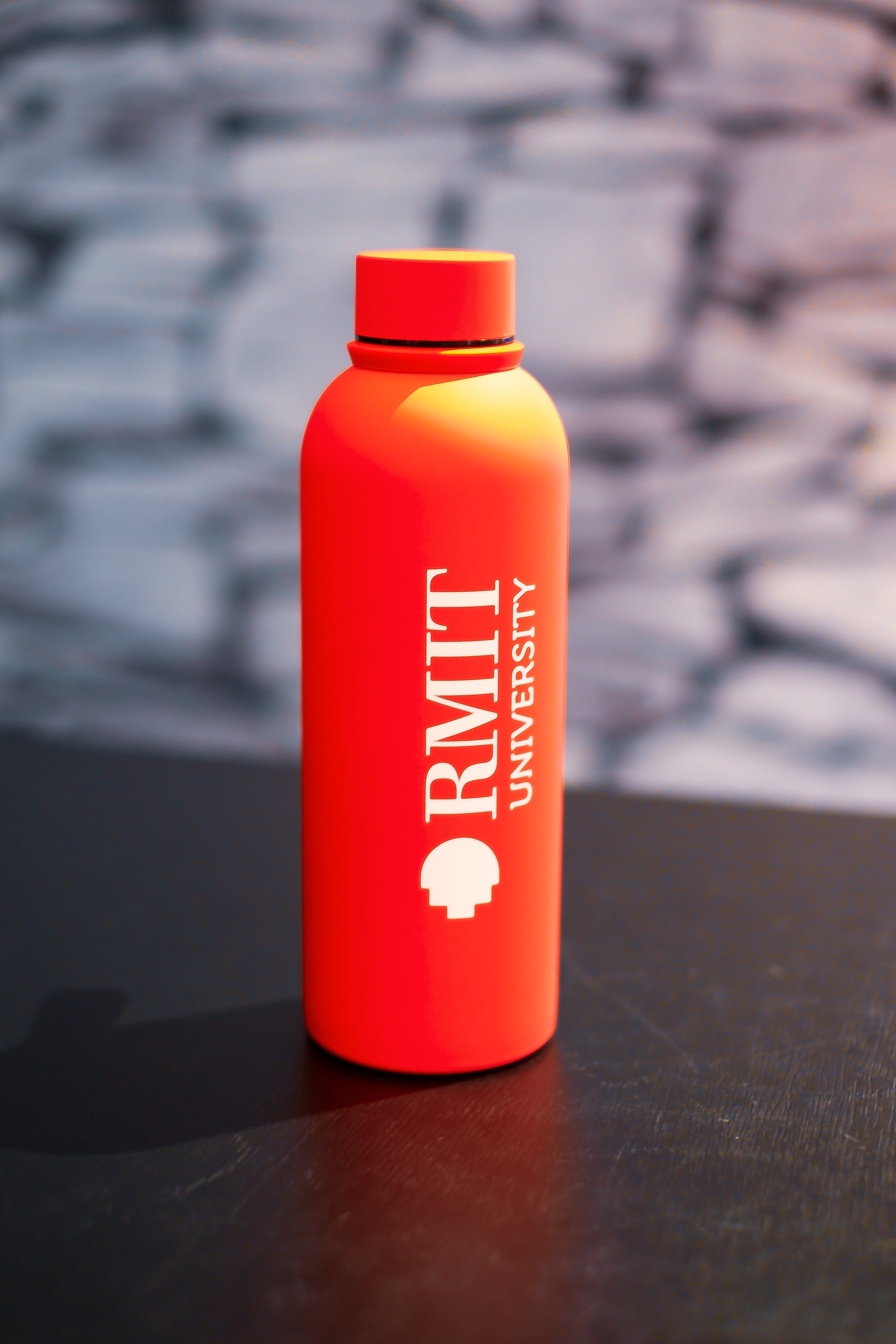 RMIT Everyday Bottle in bright red with matching screw-top lid, photographed upright on a black surface at RMIT Store, featuring vertically printed white RMIT University logo and shield icon, smooth matte coating, rounded shoulder design, and softly blurred stone-pattern background.