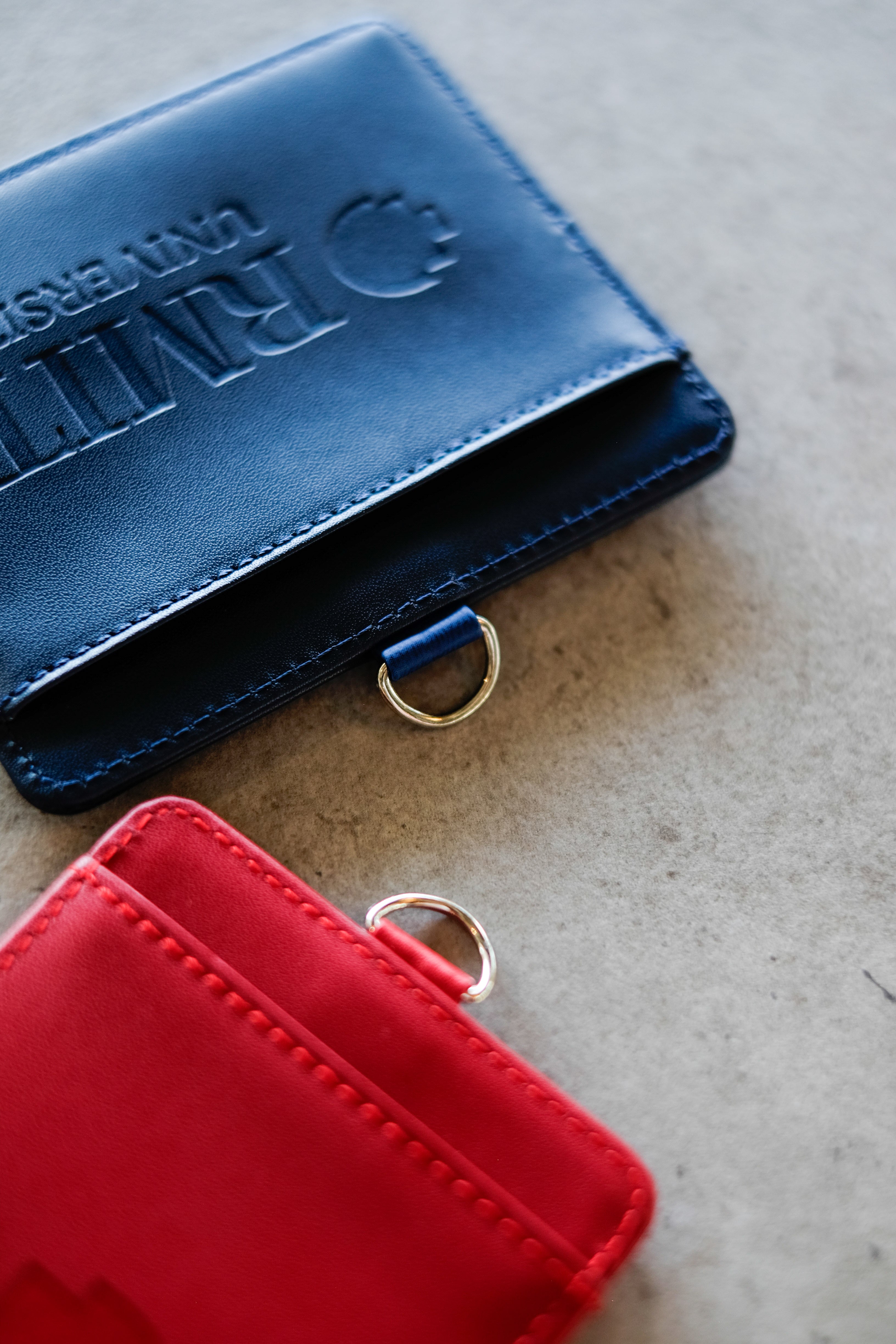 Macro close-up of the RMIT Everyday ID Card Holders showing the subtle debossed RMIT University logo and text on smooth faux leather, fine edge stitching, and the attached silver metal D-ring, photographed on a light grey surface for the RMIT Store.
