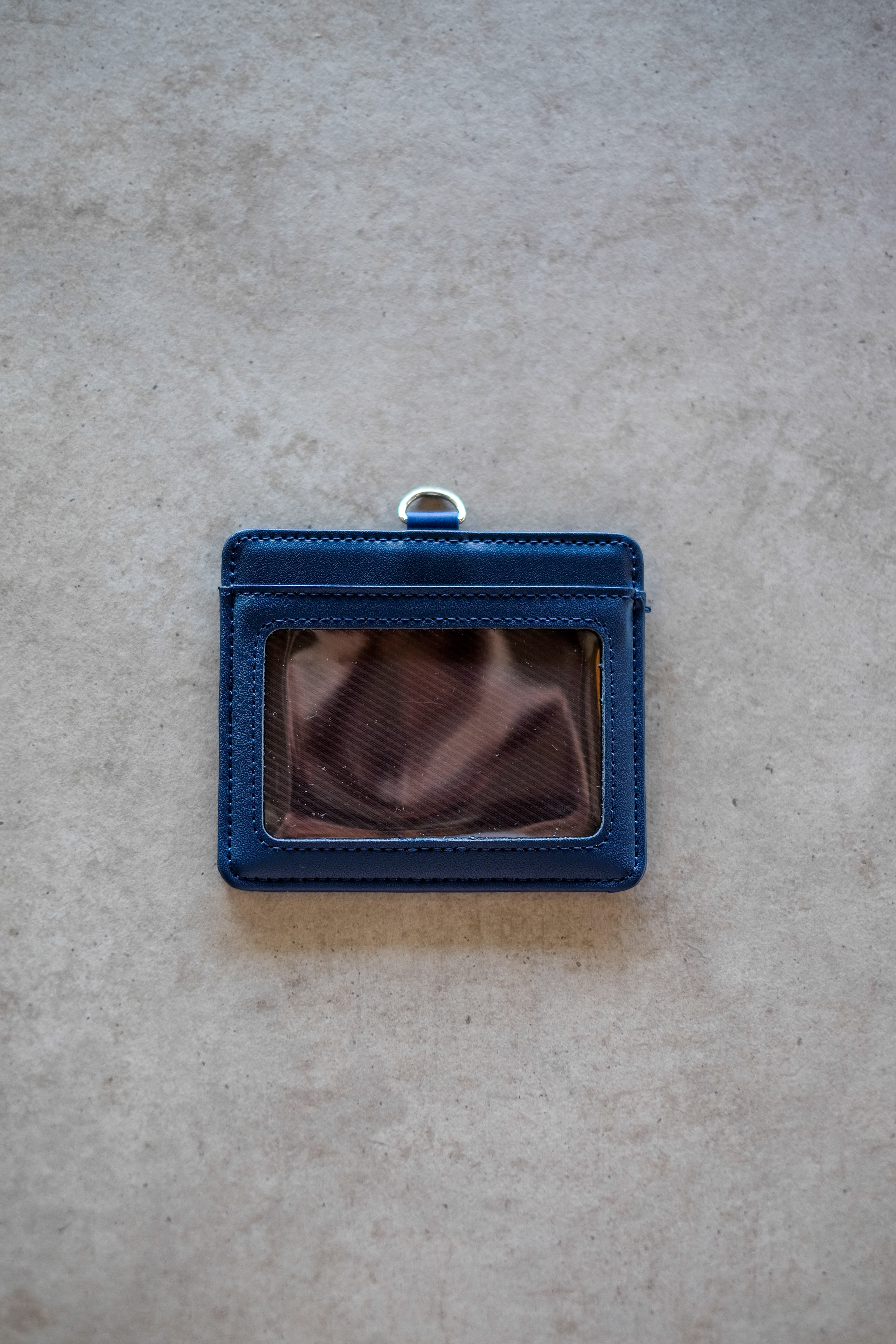 Navy RMIT Everyday ID Card Holder positioned centrally on a light grey surface, showing the front transparent ID window panel, clean stitched border, smooth faux leather texture, and a silver metal D-ring attachment at the top, photographed for the RMIT Store.