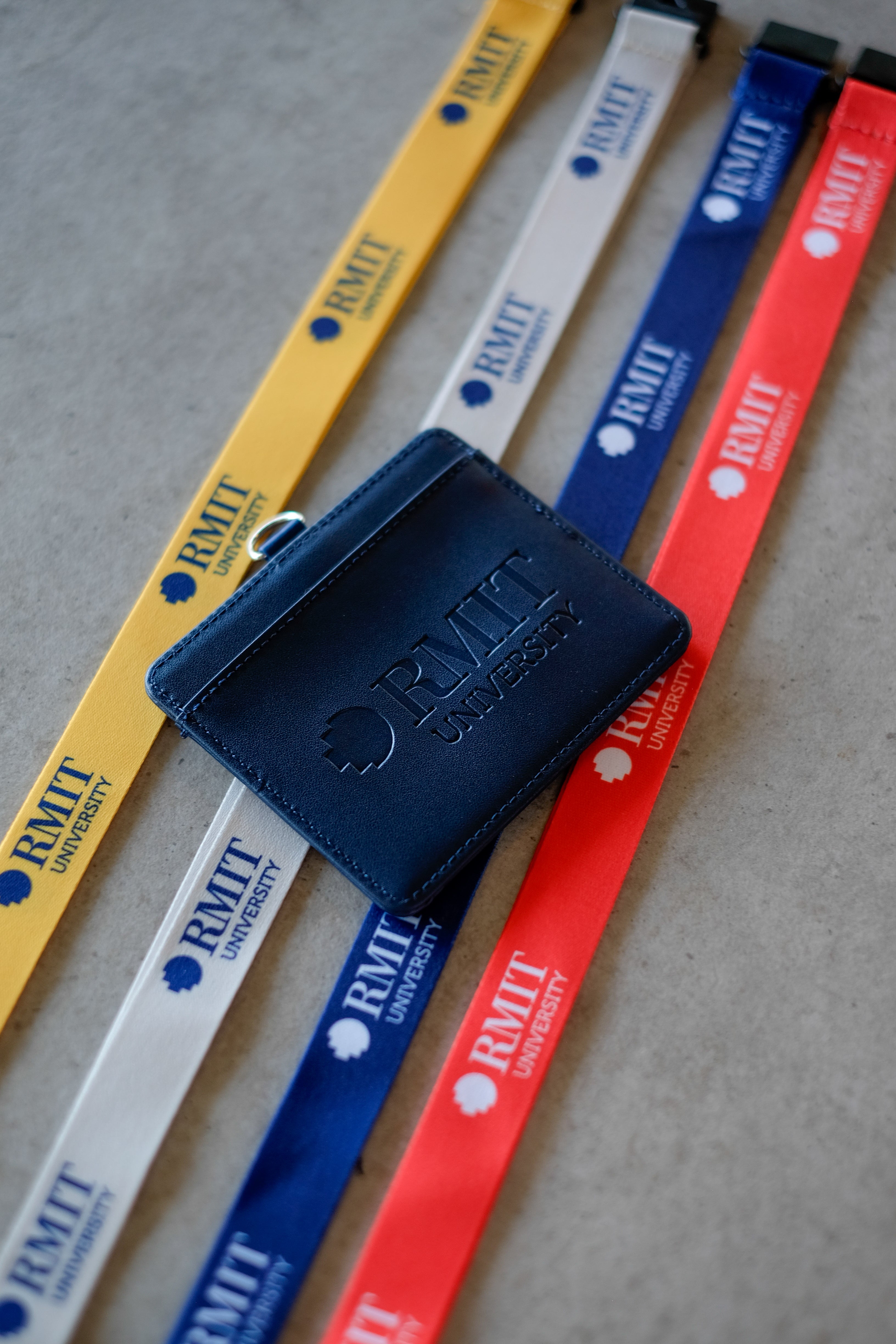 Navy RMIT Everyday ID Card Holder rests diagonally across yellow, grey, blue and red RMIT lanyards on a light grey surface, showing a subtle debossed RMIT University logo and text on the front panel, smooth faux leather finish, and a small silver metal D-ring attachment, photographed for the RMIT Store.