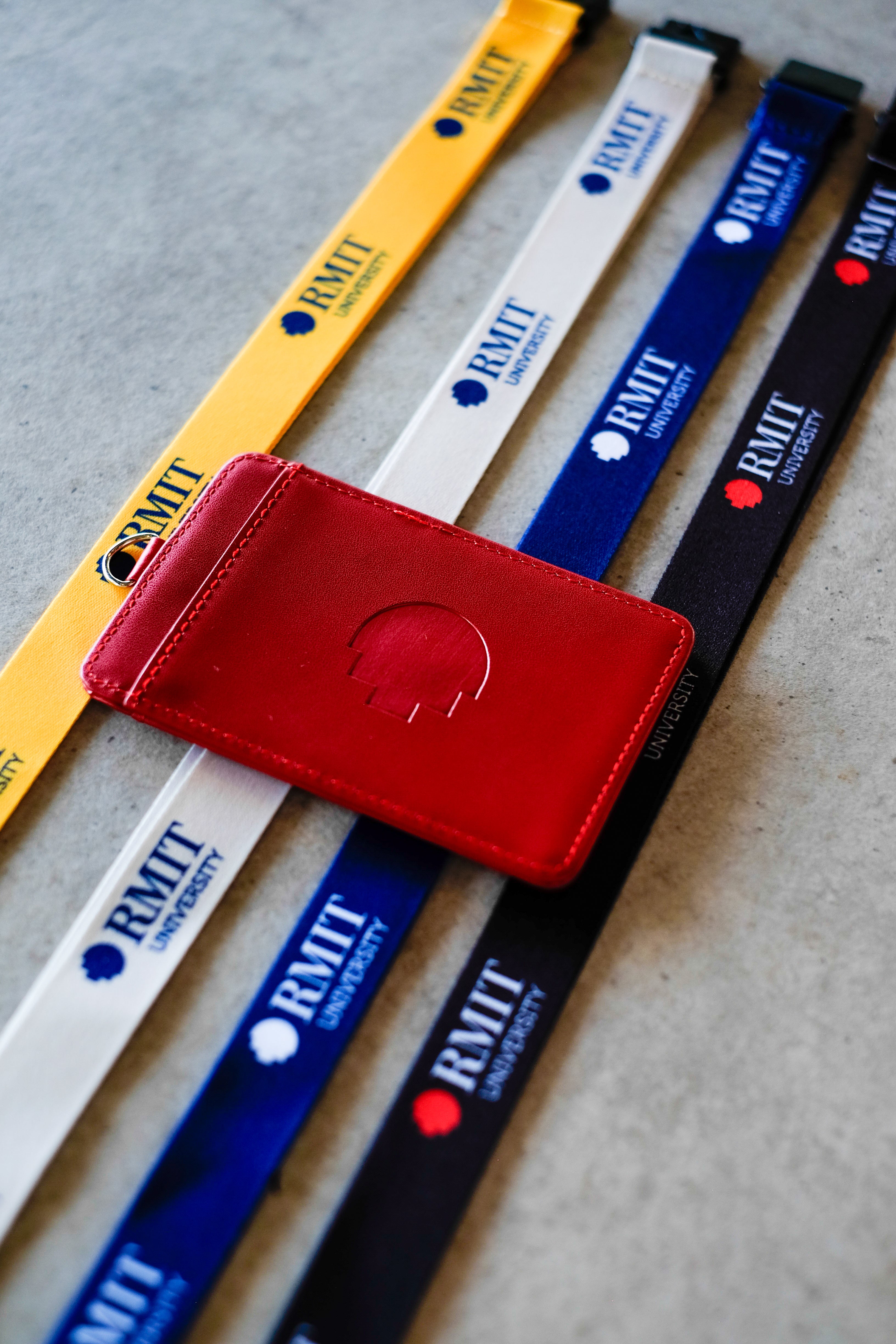 Red RMIT Everyday ID Card Holder made from smooth faux leather lies diagonally across yellow, grey, navy and black RMIT lanyards on a light grey surface, showing an embossed RMIT University shield logo centred on the front panel, neat edge stitching, and a small silver metal D-ring attachment, photographed for the RMIT Store.