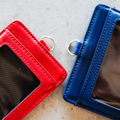 Close-up detail of red and navy RMIT Everyday ID Card Holders placed side by side on a light grey surface, showing precise stitching, smooth faux leather texture, curved corner edges, and small silver metal D-ring attachments, photographed for the RMIT Store.