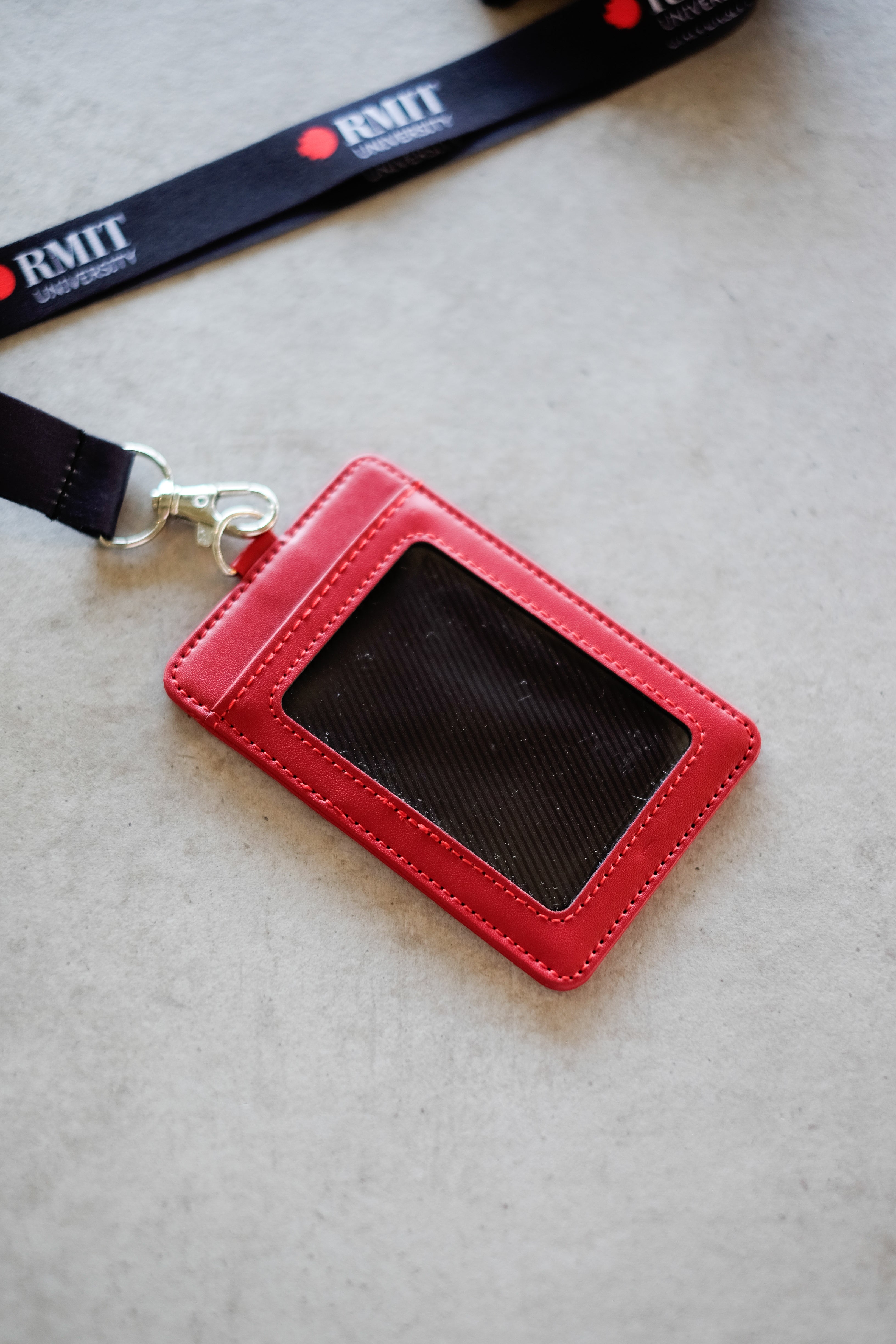 Red RMIT Everyday ID Card Holder displayed flat on a light grey surface, showing the rear transparent ID window panel with visible stitching, smooth faux leather edges, and a silver metal lobster clasp connected to a black RMIT lanyard, photographed for the RMIT Store.