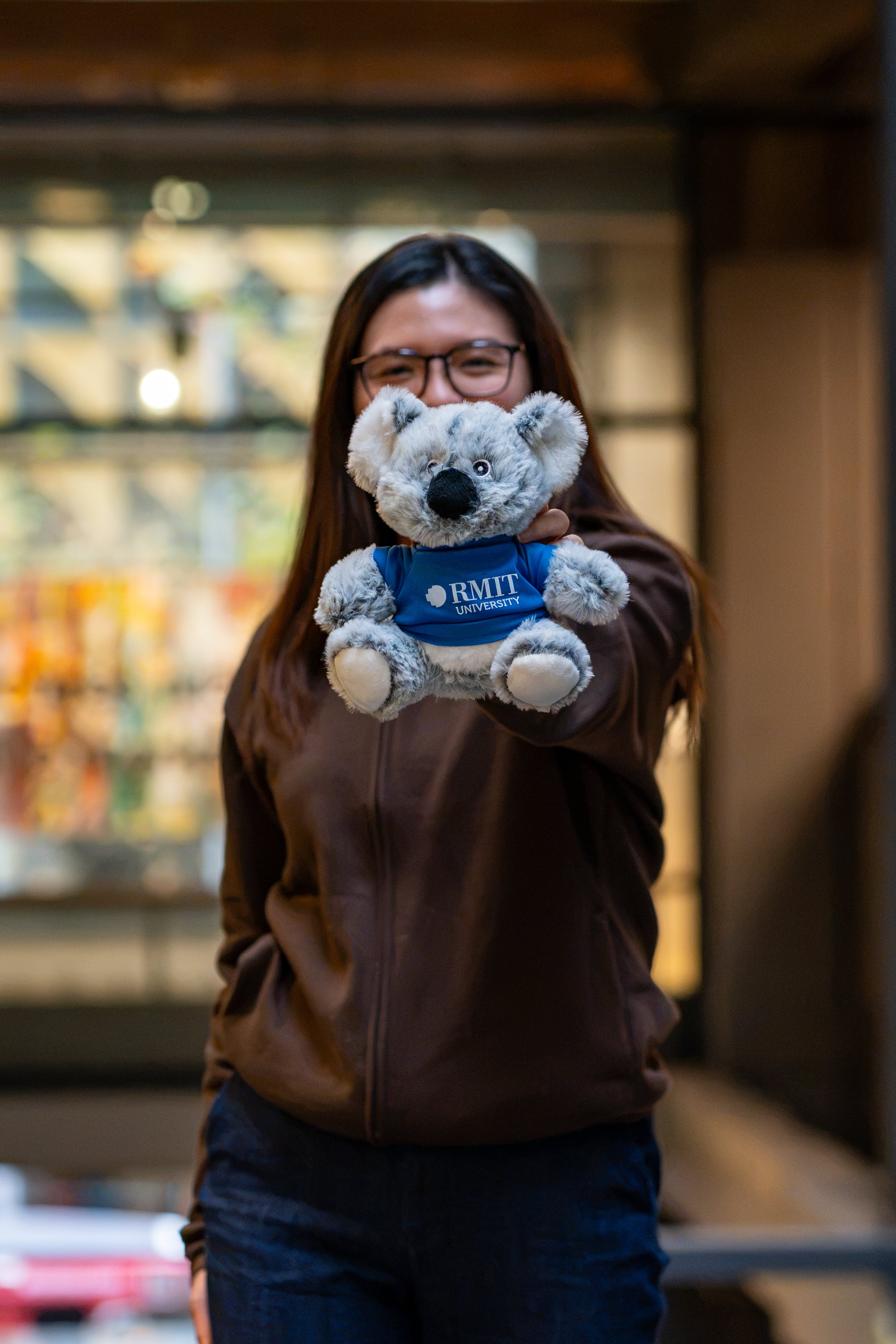 RMIT Store RMIT Graduation Koala plush with soft grey fur wearing a dark blue T-shirt featuring a white RMIT University logo on the front, extended towards the camera by a model wearing a chocolate RMIT University zip jacket, highlighting the embroidered logo and plush details against a blurred campus background.