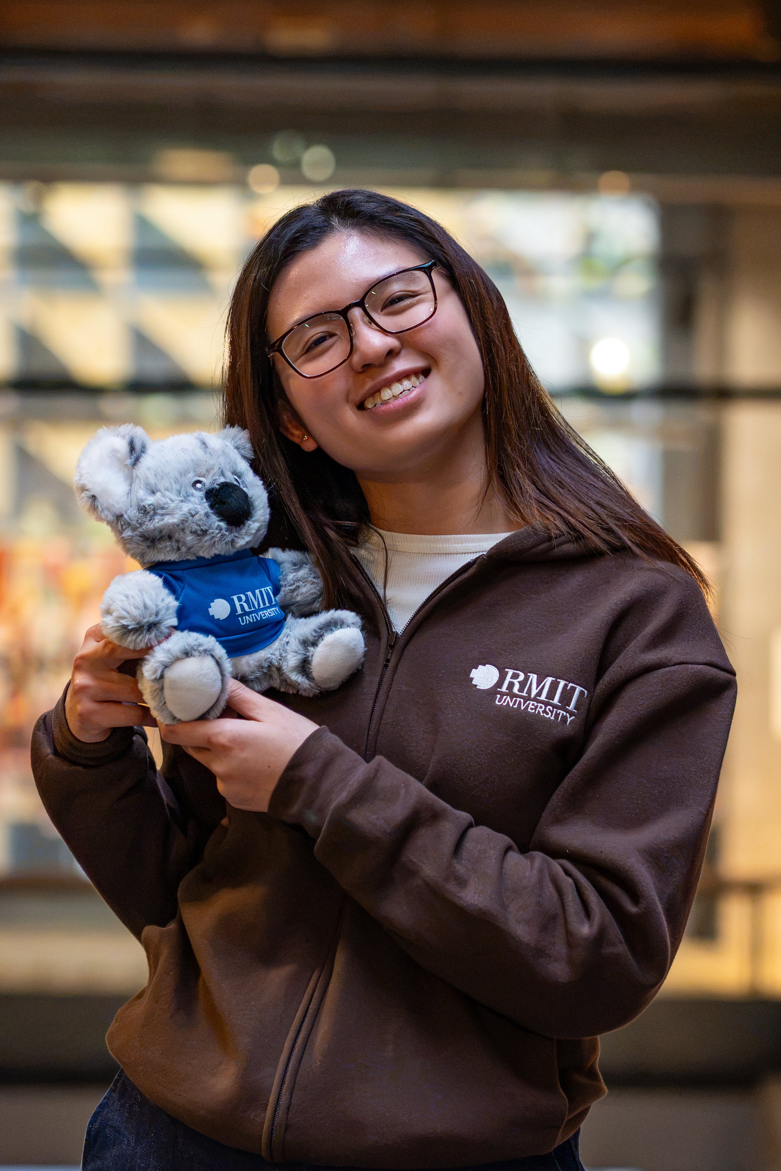 RMIT Graduation Koala plush with soft grey fur wearing a dark blue T-shirt featuring a white RMIT University logo on the front, held by a smiling model wearing a chocolate RMIT University zip jacket with a white embroidered chest logo in an outdoor campus setting with a softly blurred background.