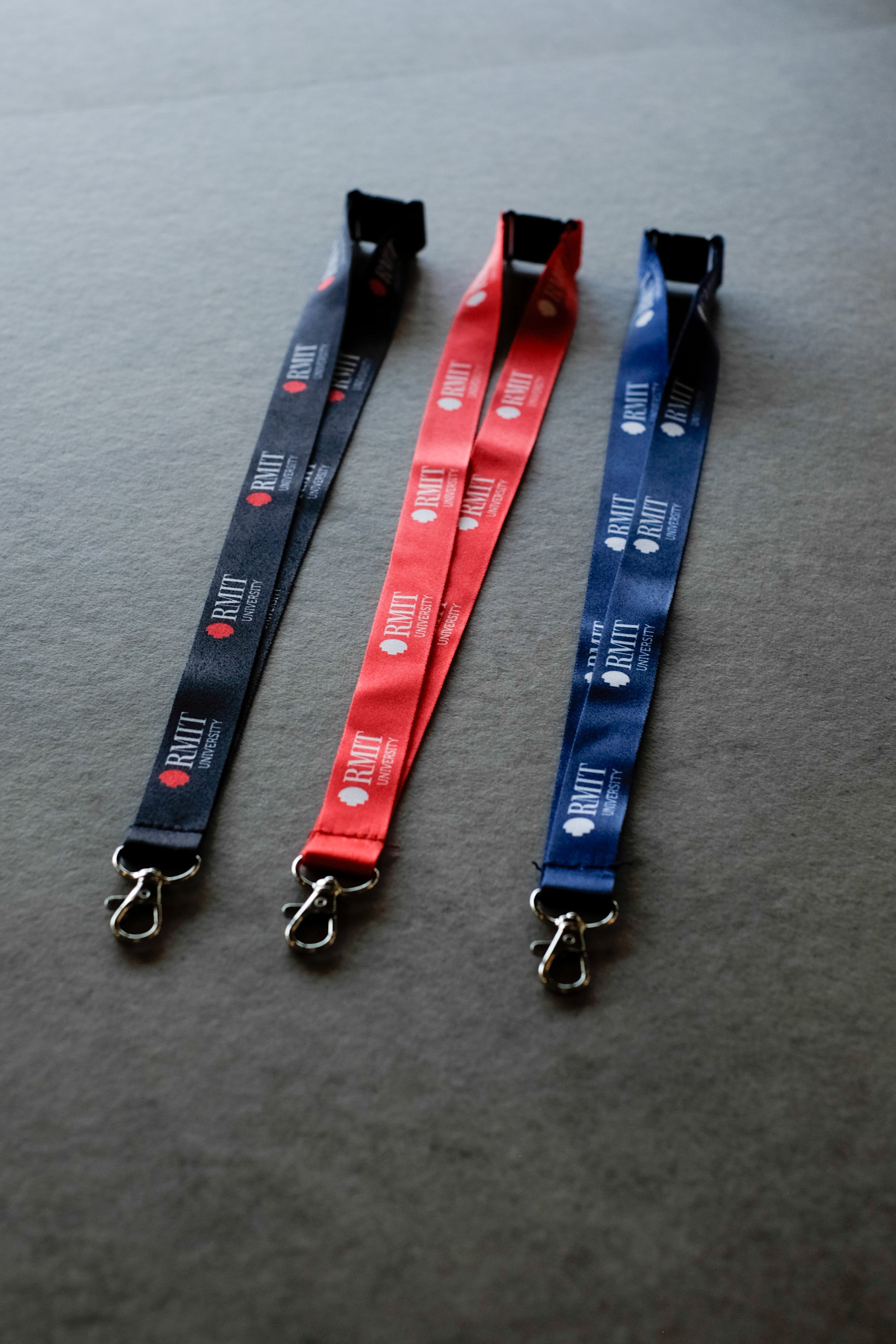 Three RMIT Lanyards in black, red, and navy blue lie flat side-by-side on a light grey fabric surface, each featuring repeated white RMIT University logo prints along the woven polyester strap, a black plastic safety breakaway clip at the top, and a silver metal swivel lobster clasp at the end, as shown in this product image available from the RMIT Store.