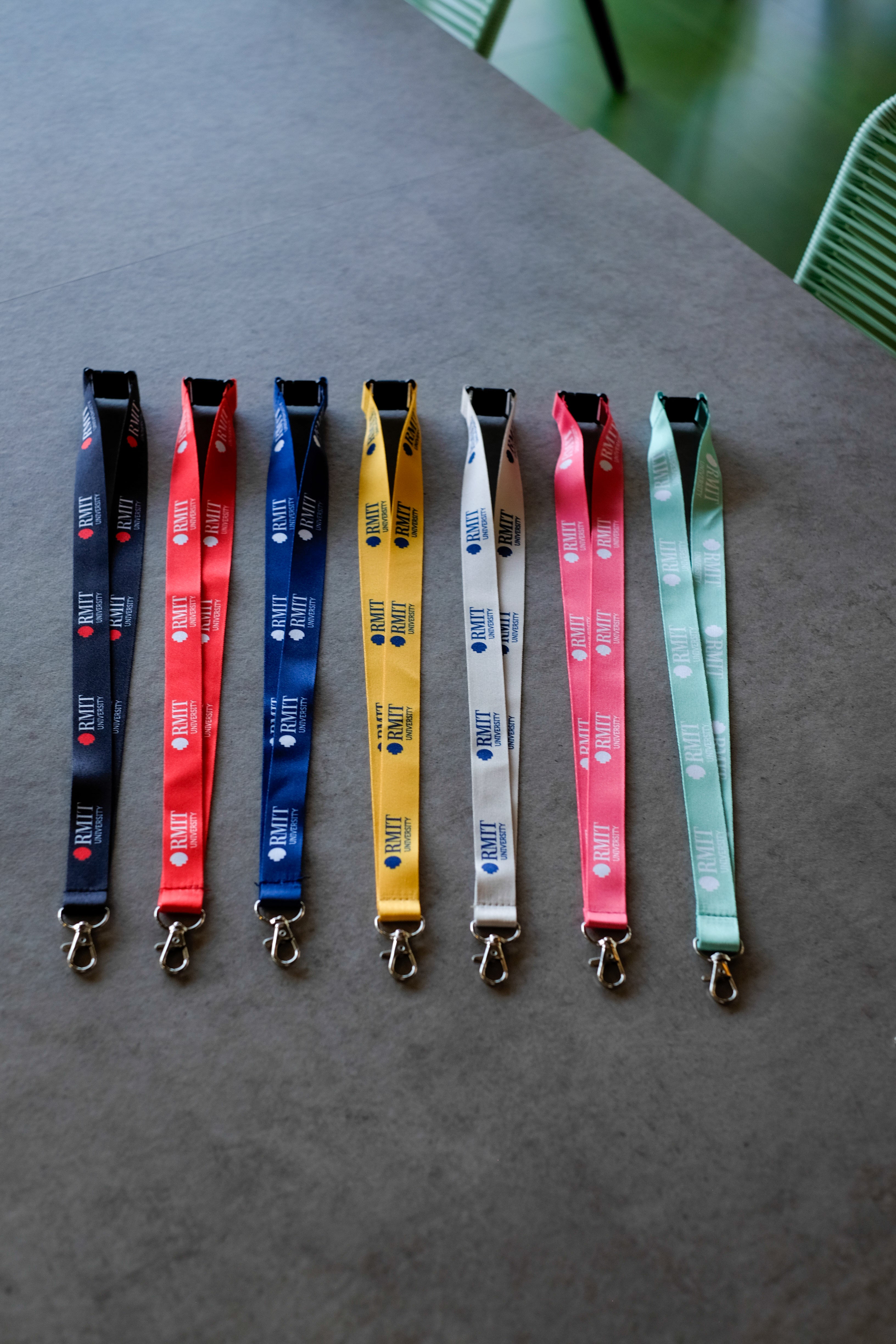 Seven RMIT Lanyards in black, red, blue, yellow, grey, pink, and green are arranged side by side on a light grey tabletop, each showing repeated RMIT University logo prints along the woven polyester strap, a black plastic safety breakaway clip at the top, and a silver metal swivel lobster clasp at the end, with green chair legs softly visible in the background in this product image from the RMIT Store.