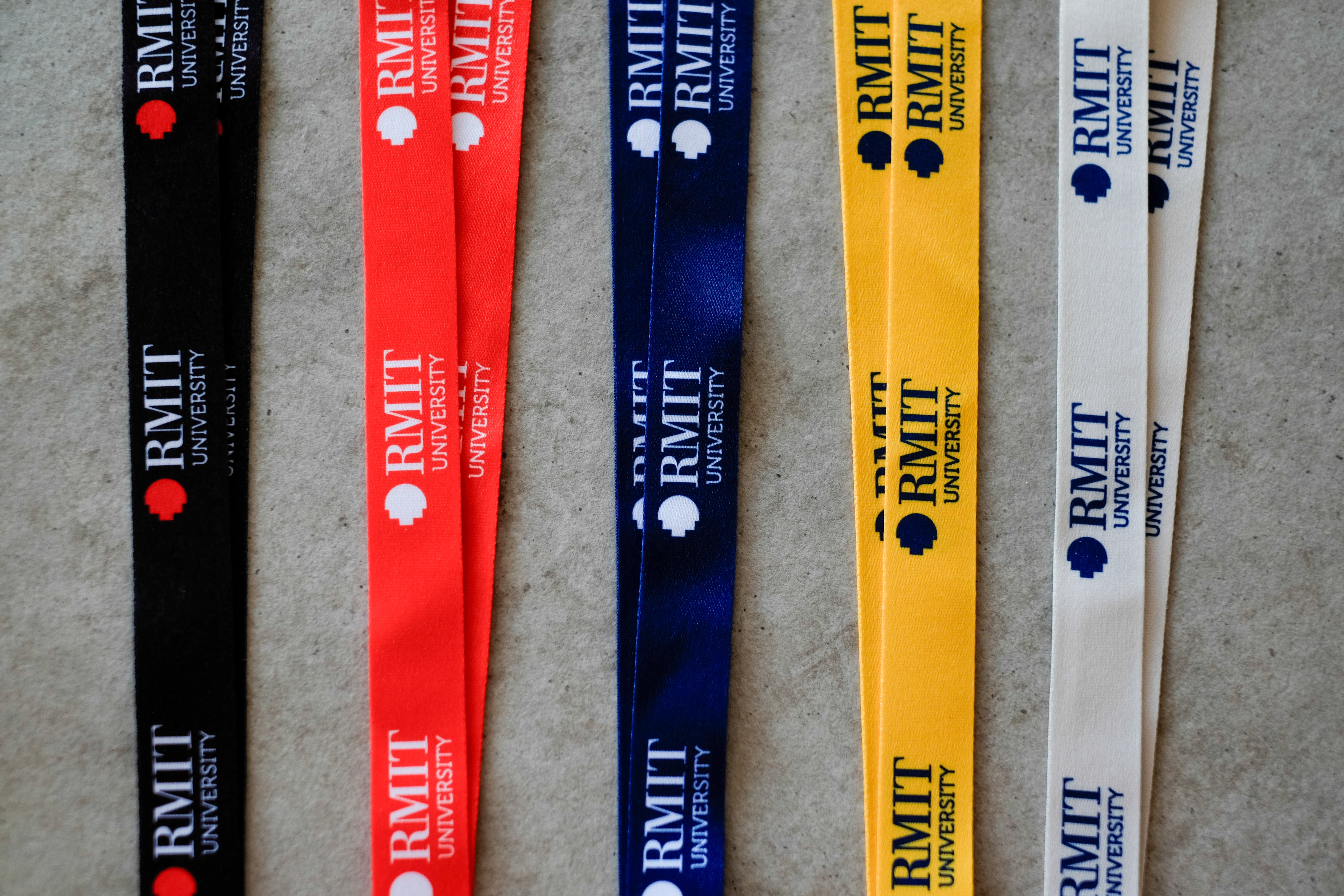 Close-up of five RMIT Lanyards in black, red, navy blue, yellow and grey laid flat on a light grey surface, showing repeated woven RMIT University logo and text prints along each polyester strap, with the fabric texture and crisp logo detail clearly visible in this product image from the RMIT Store.
