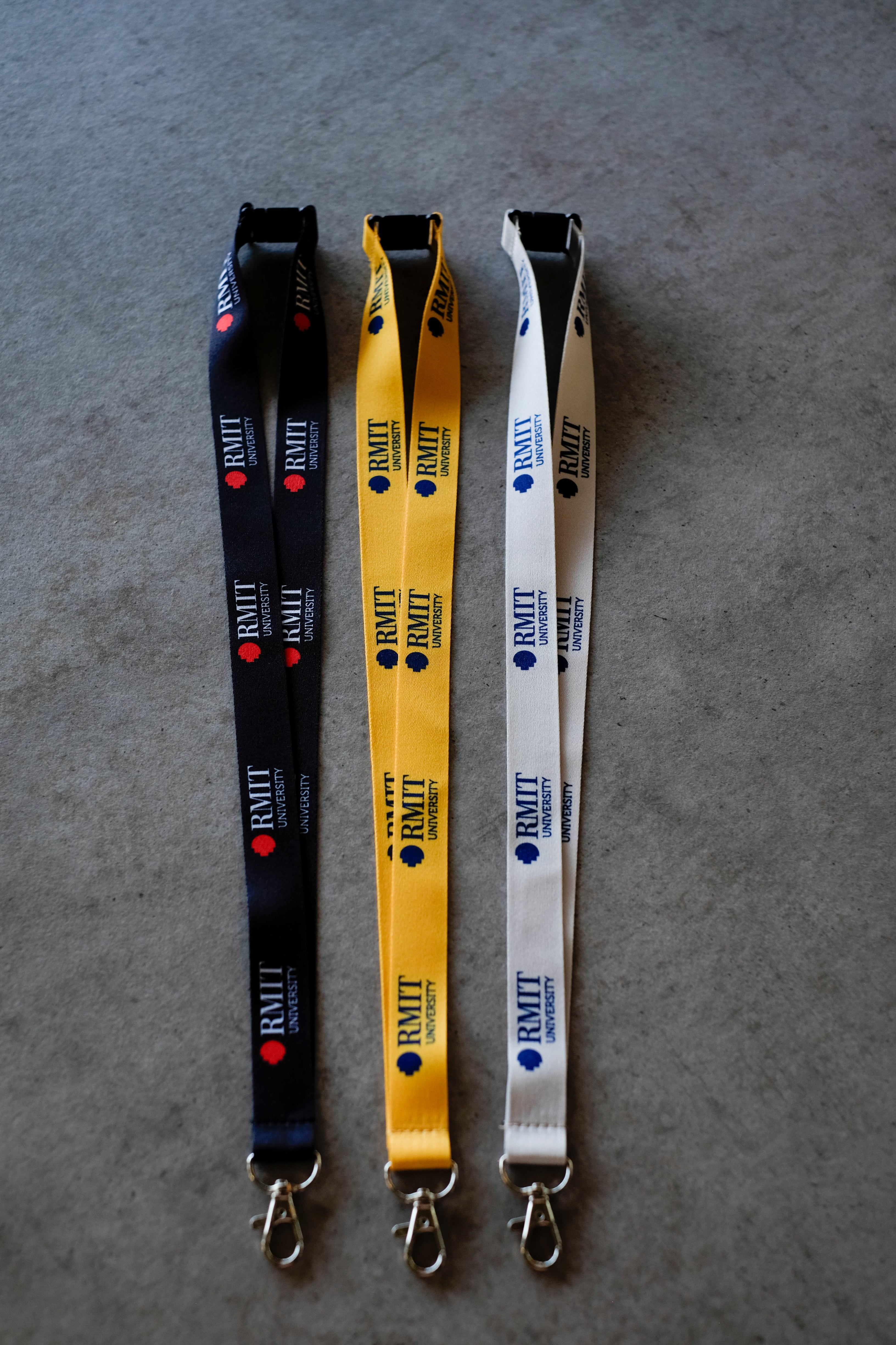 Three RMIT Lanyards in black, yellow and grey are laid flat side by side on a light grey fabric surface, each featuring repeated woven RMIT University logos along the strap, a black plastic safety breakaway clip at the top, and a silver metal swivel lobster clasp at the end, as shown in this product image available from the RMIT Store.