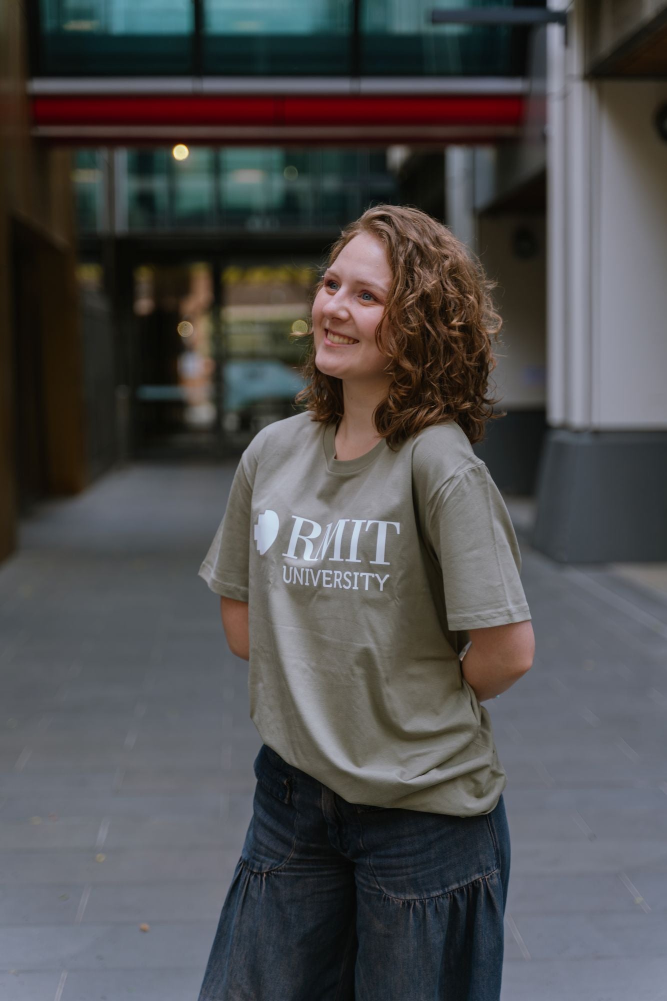 Model standing in an outdoor campus walkway wearing RMIT Loud Logo Tee ’26 in eucalyptus green featuring a large white RMIT University logo printed across the chest, styled with dark denim wide-leg jeans, smiling with hands behind back and head slightly turned, with blurred modern building background, available at the RMIT Store.