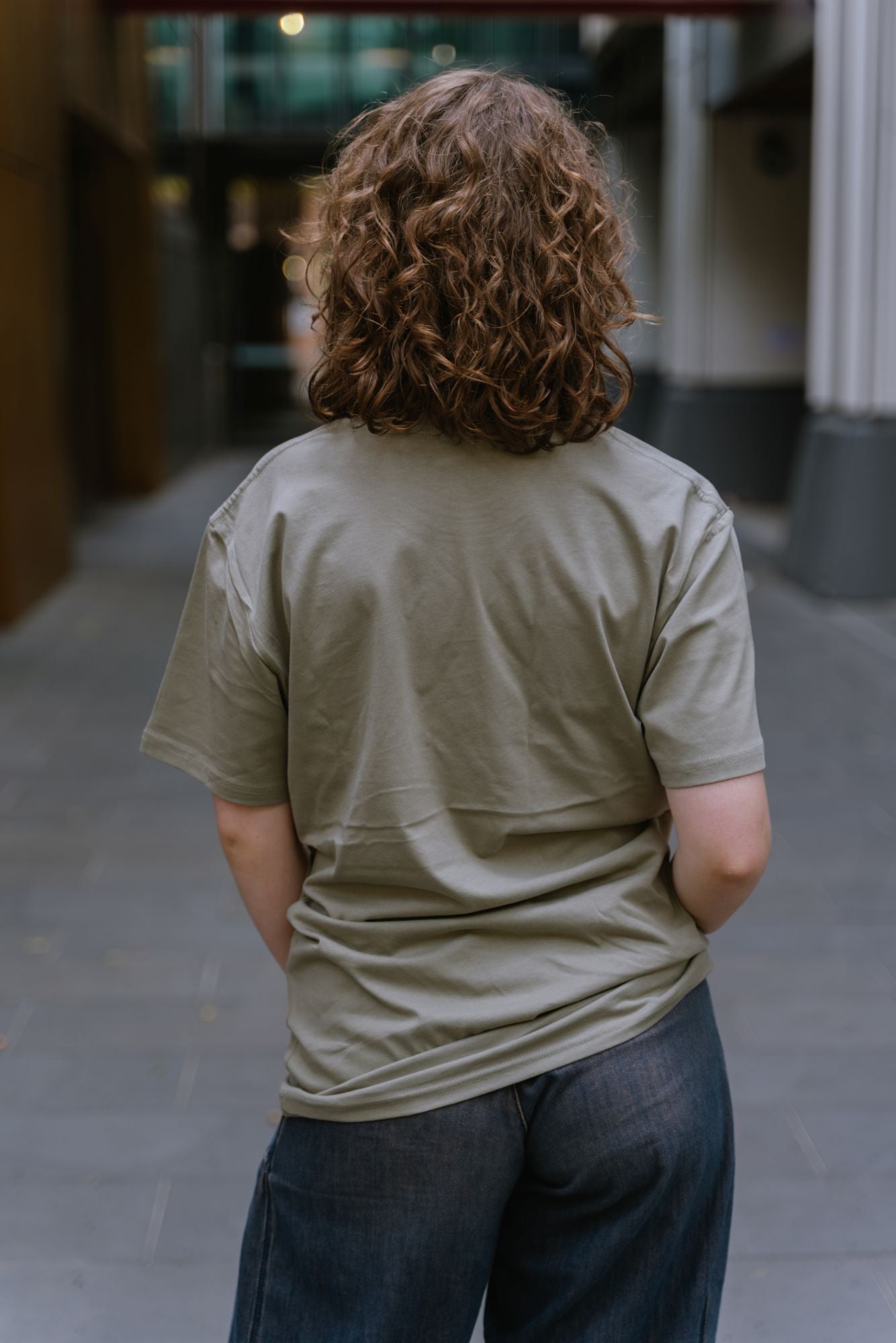 Rear view of model standing in outdoor campus walkway wearing RMIT Loud Logo Tee ’26 in eucalyptus green, showing plain back with no visible print and soft fabric drape, paired with dark denim wide-leg jeans, with blurred modern building background, available at the RMIT Store.