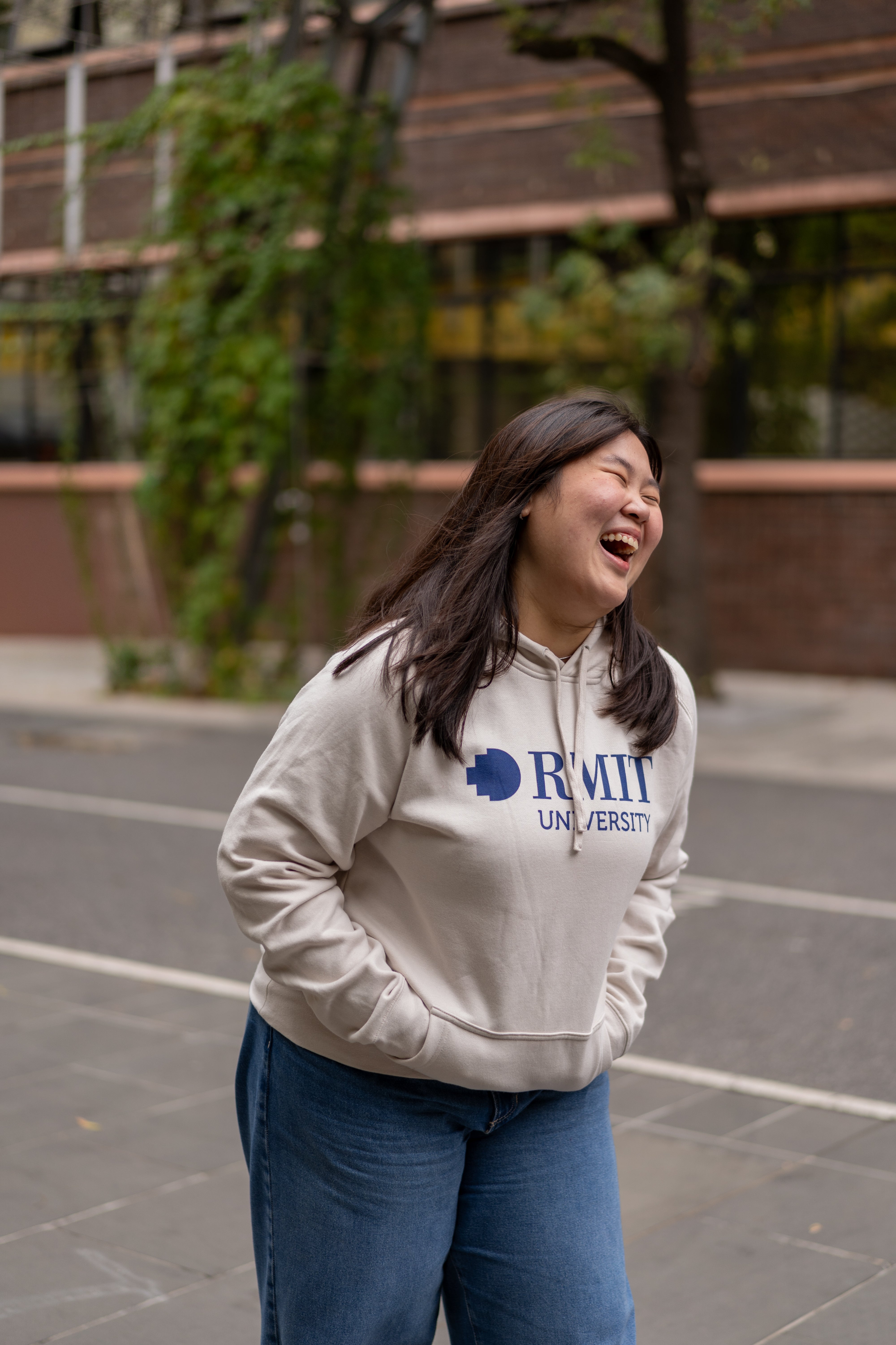 Smiling model in RMIT Store Limited Edition Bone Hoodie, showcasing the relaxed fit, soft bone-coloured cotton material, and large navy RMIT University logo printed across the chest.