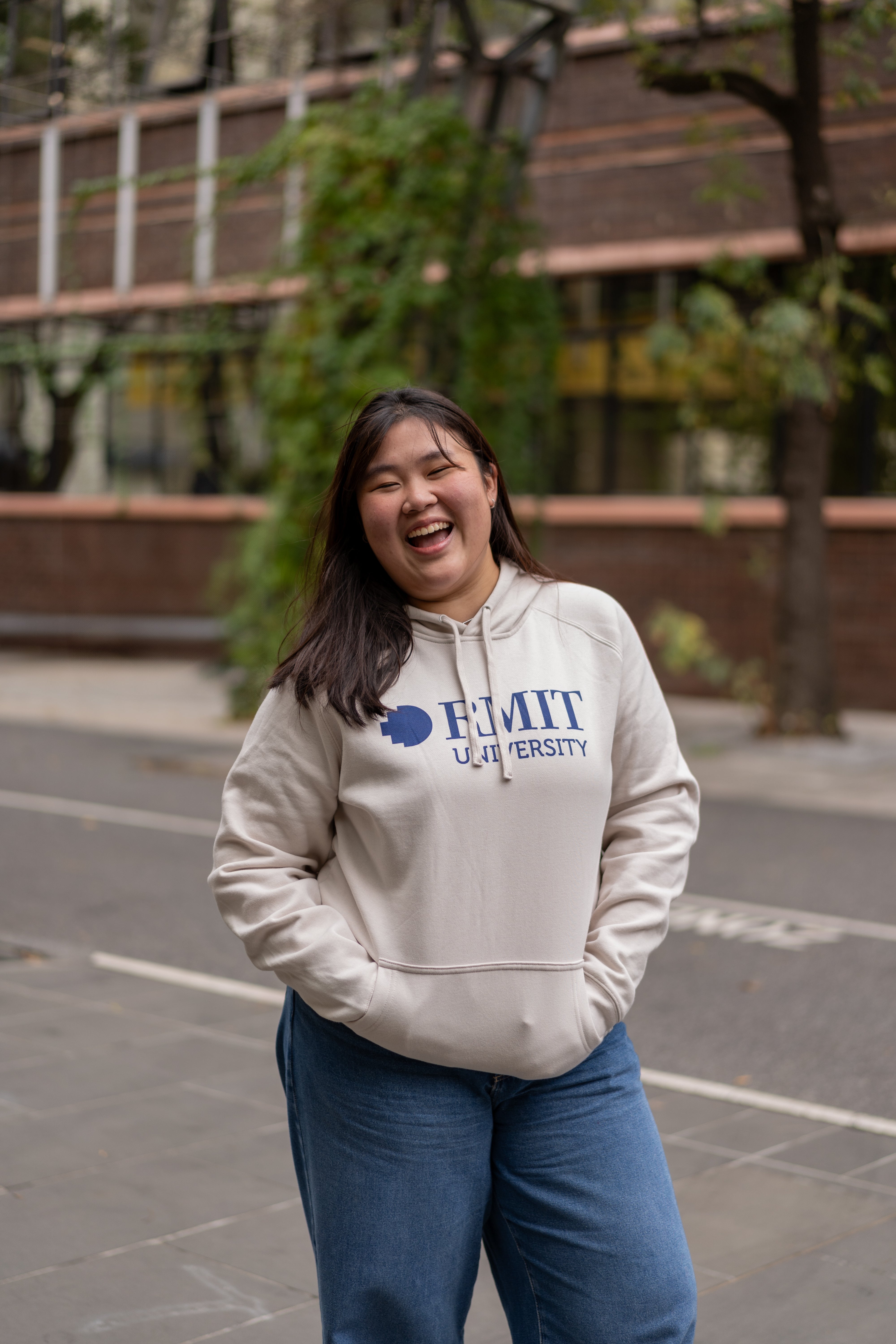 Model wearing the RMIT Store Limited Edition Bone Hoodie with hands in the pocket, featuring the bold RMIT University logo in navy blue, front pocket design, and adjustable drawstring hood, styled in an outdoor urban setting.