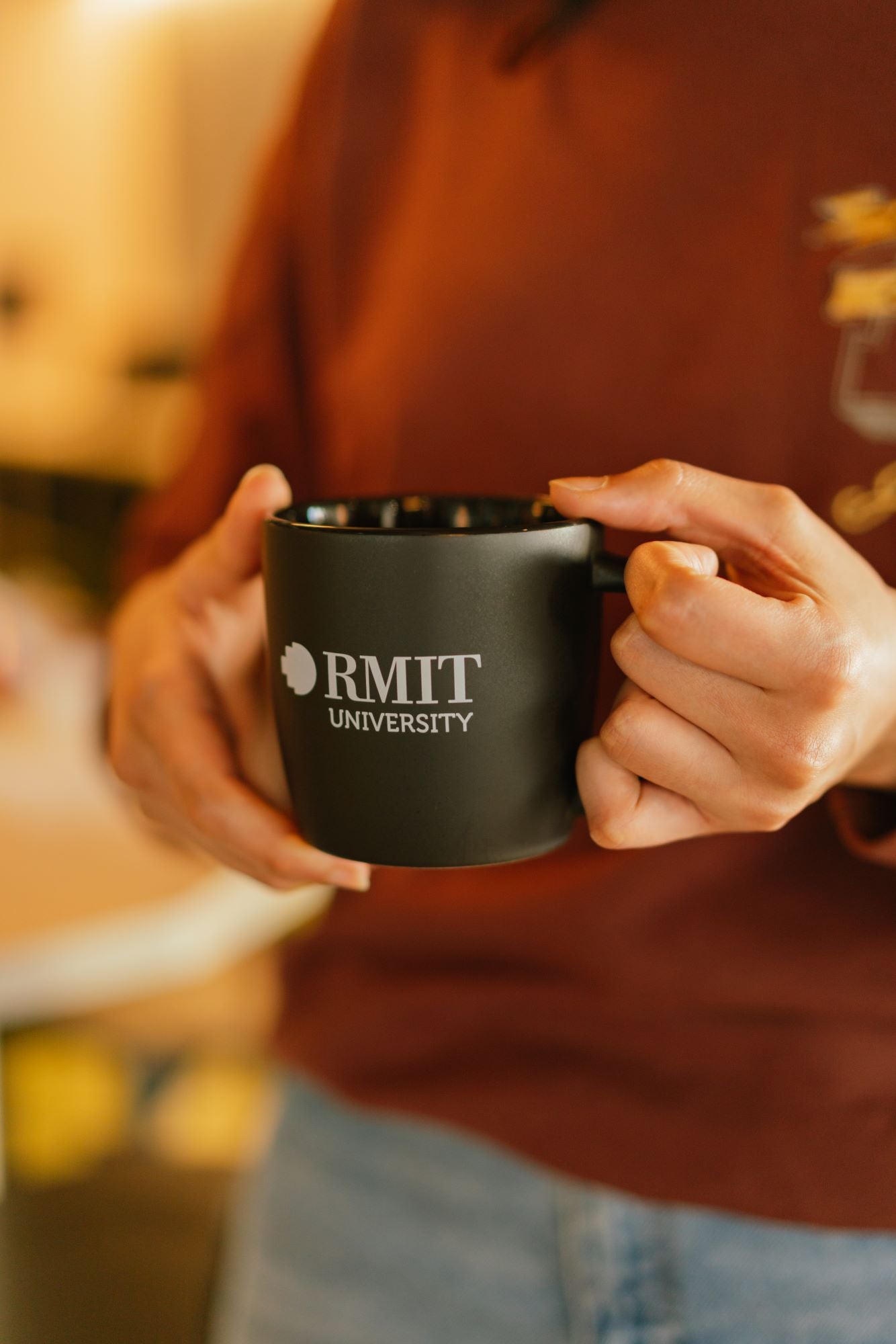 Model holding a black ceramic RMIT Store mug with black interior in both hands, showing the white RMIT University logo on the front. 