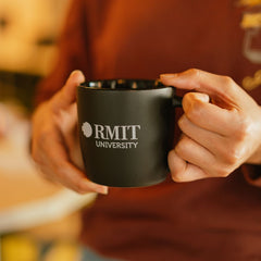 Model holding a black ceramic RMIT Store mug with black interior in both hands, showing the white RMIT University logo on the front. 