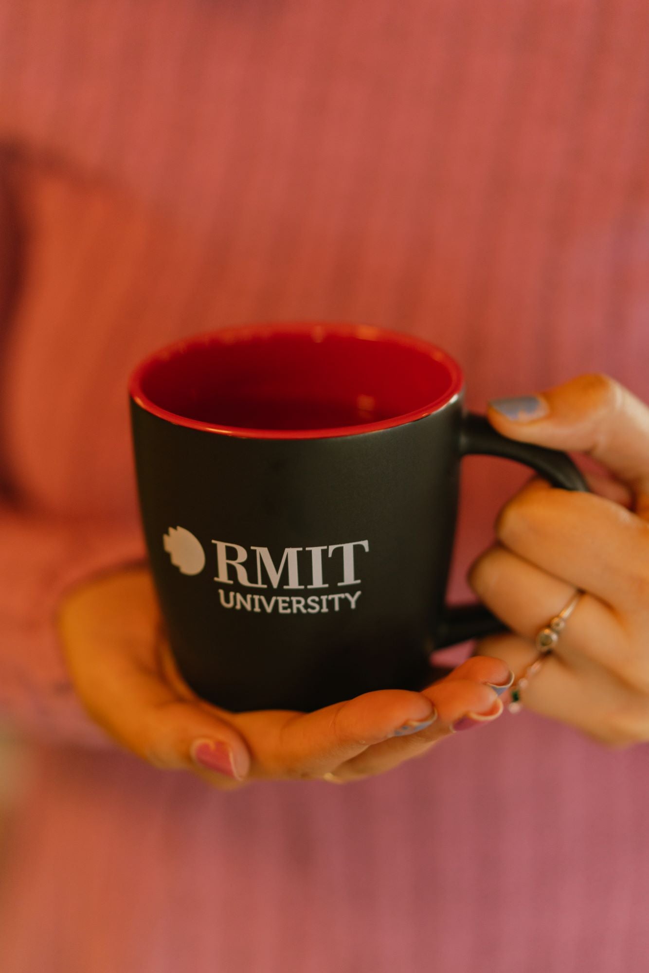 Model holding a black ceramic RMIT Store mug with a red interior and white RMIT University logo printed on the outer surface, positioned at chest level with the handle facing right, against a softly blurred neutral background.