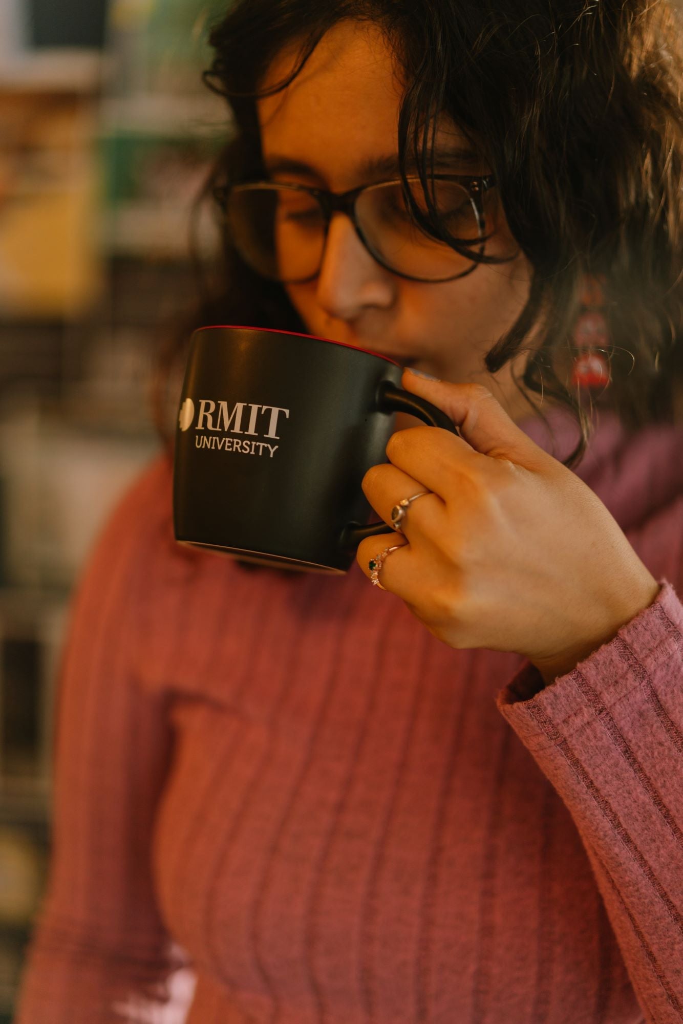 Model wearing a pink sweater drinking from a black ceramic RMIT Store mug with red interior, holding it in her left hand. The mug features a white RMIT University logo on the front. 