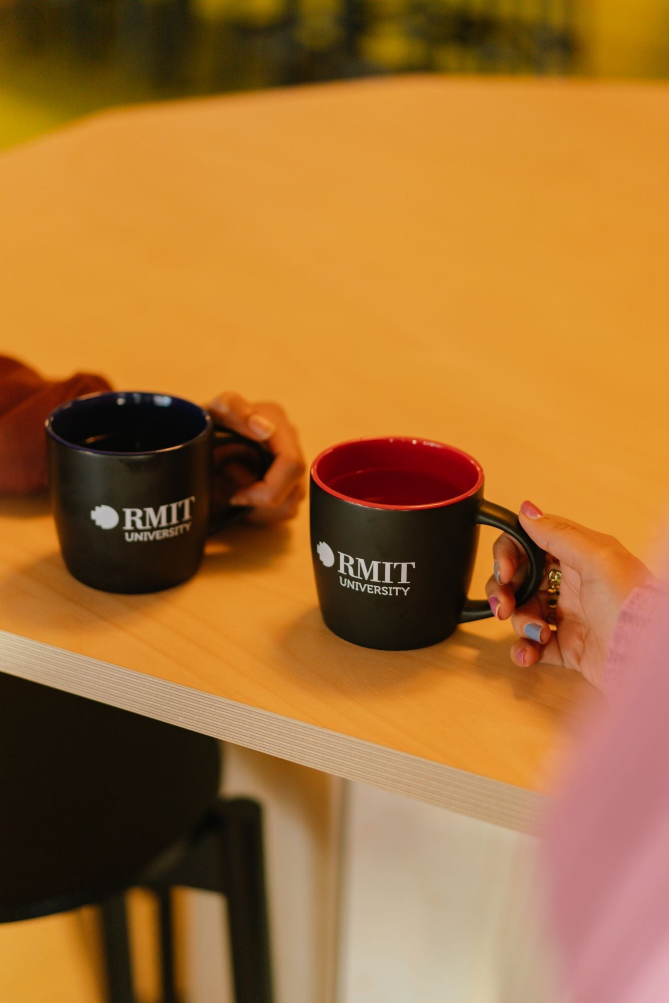 Close-up of two models’ hands holding different RMIT Store mugs above a wooden tabletop; the mug on the left is black ceramic with black interior, and the mug on the right is black ceramic with red interior. Both mugs display a white RMIT University logo on the front.