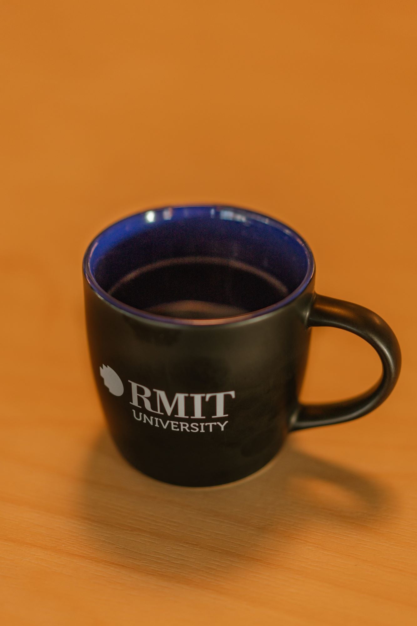 RMIT Store indigo ceramic mug with white RMIT University logo prominently printed on one side, featuring a curved handle and matte finish, displayed upright on a light wooden tabletop with soft natural lighting.