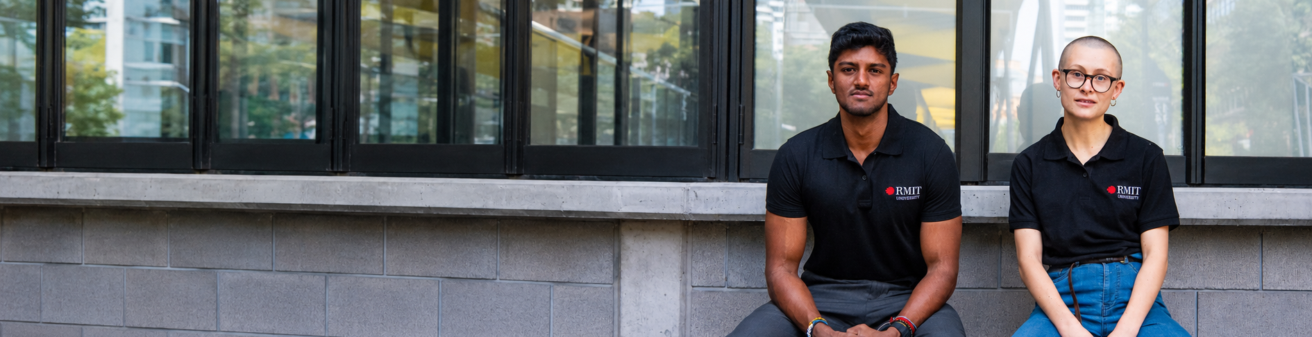 RMIT Store RMIT Polo in black, modelled from the front by two seated models, featuring short sleeves, a pointed collar, buttoned placket and embroidered red-and-white RMIT University logo on the left chest, styled with dark trousers and blue jeans outside a modern building with grey brickwork and large glass windows.