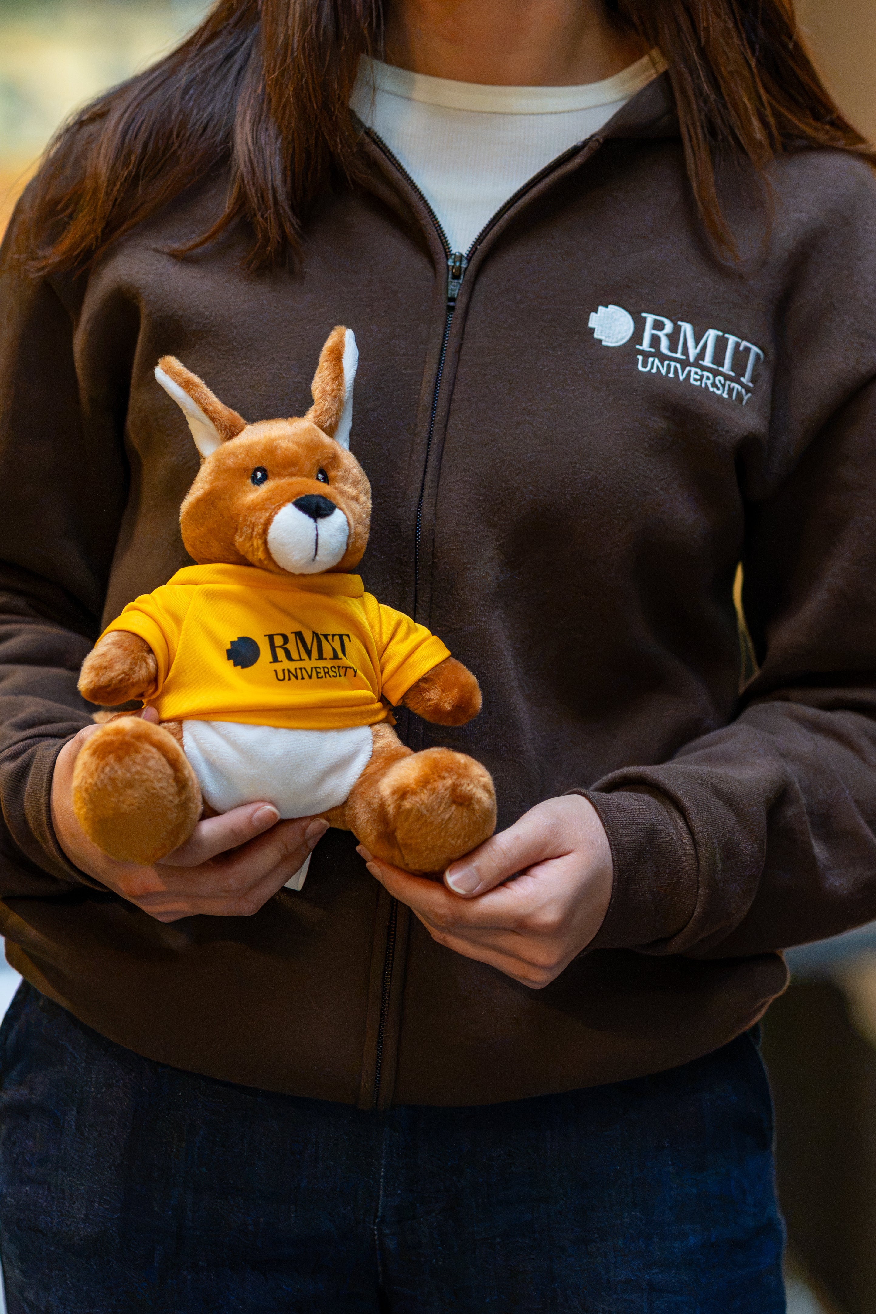 RMIT Store RMIT University Kangaroo Plush in a yellow branded tee, held front-facing by a model wearing a chocolate RMIT University zip jacket with embroidered chest logo.