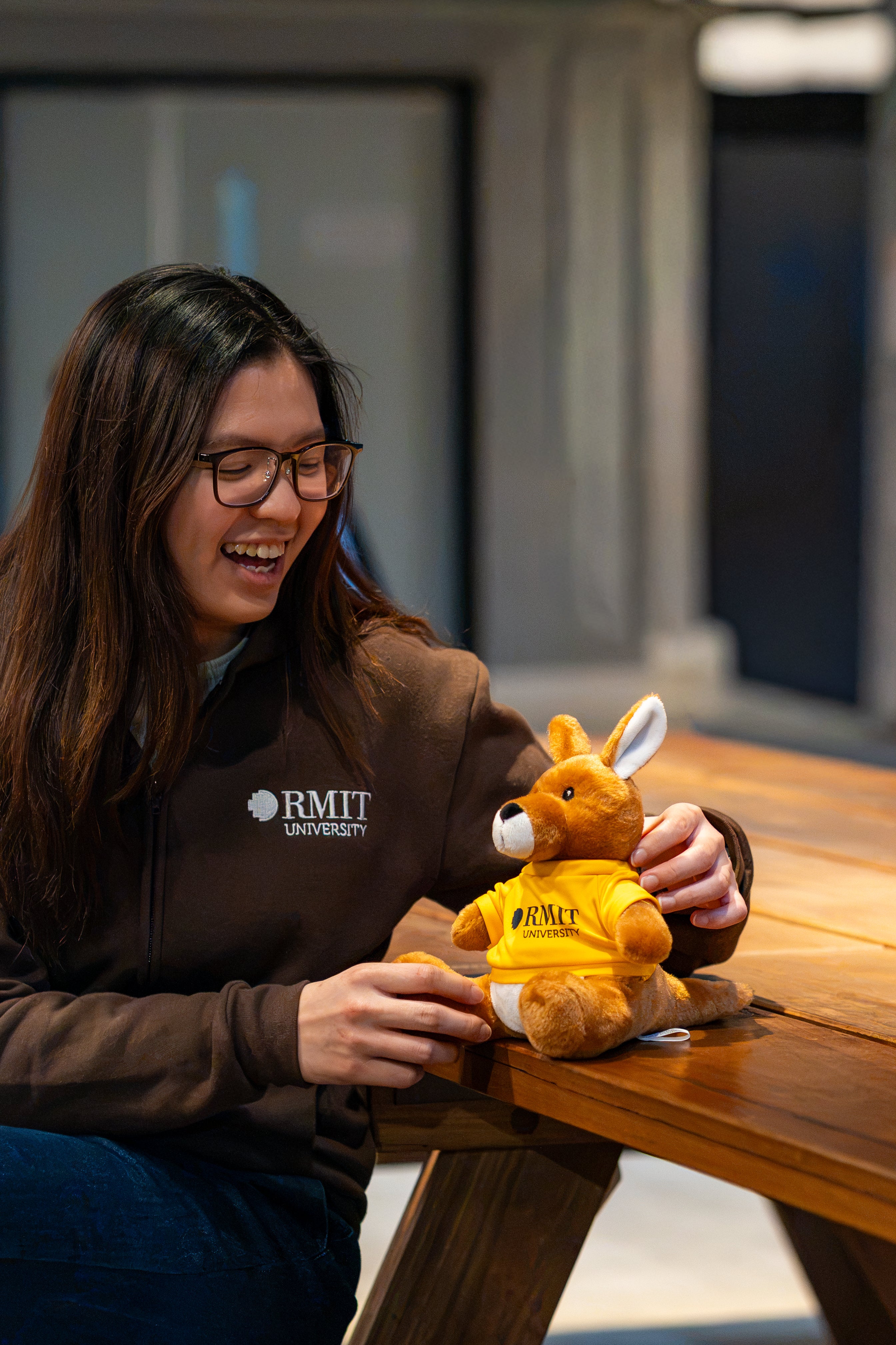 RMIT Store RMIT University Kangaroo Plush in a yellow branded tee seated on a wooden table beside a smiling model wearing a chocolate RMIT University zip jacket.