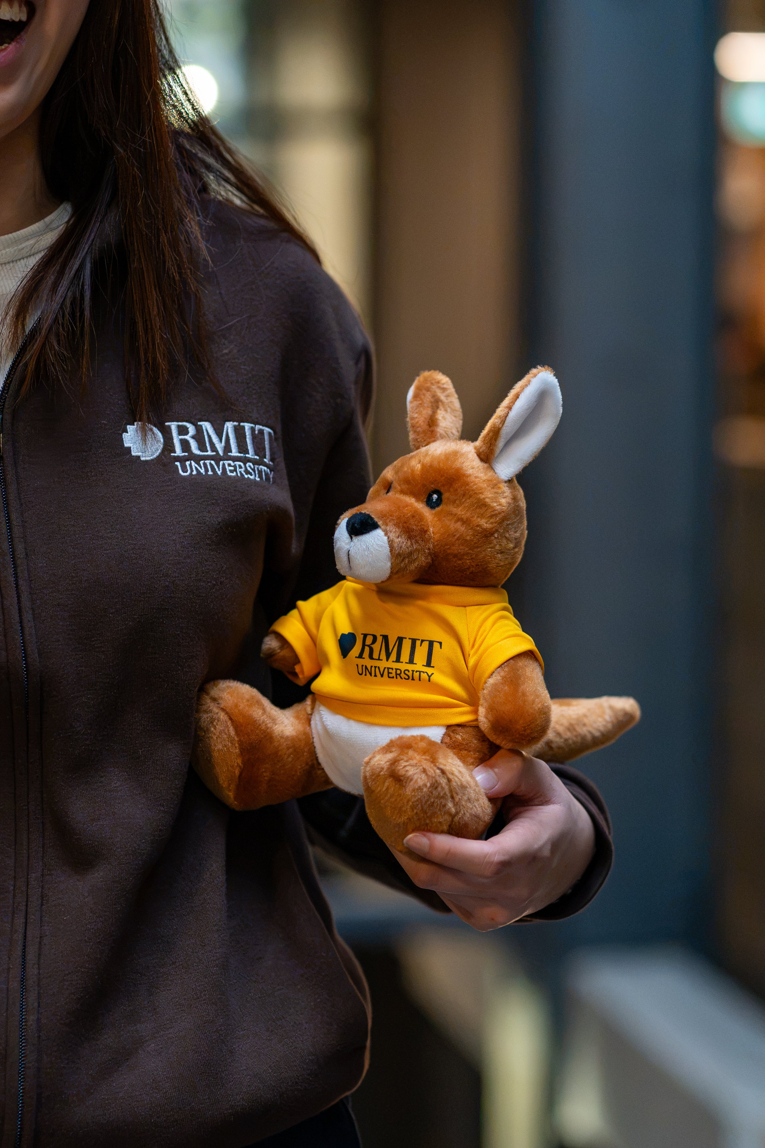 RMIT Store RMIT University Kangaroo Plush in a yellow branded tee shown from the side, held by a smiling model in a chocolate RMIT University zip jacket.