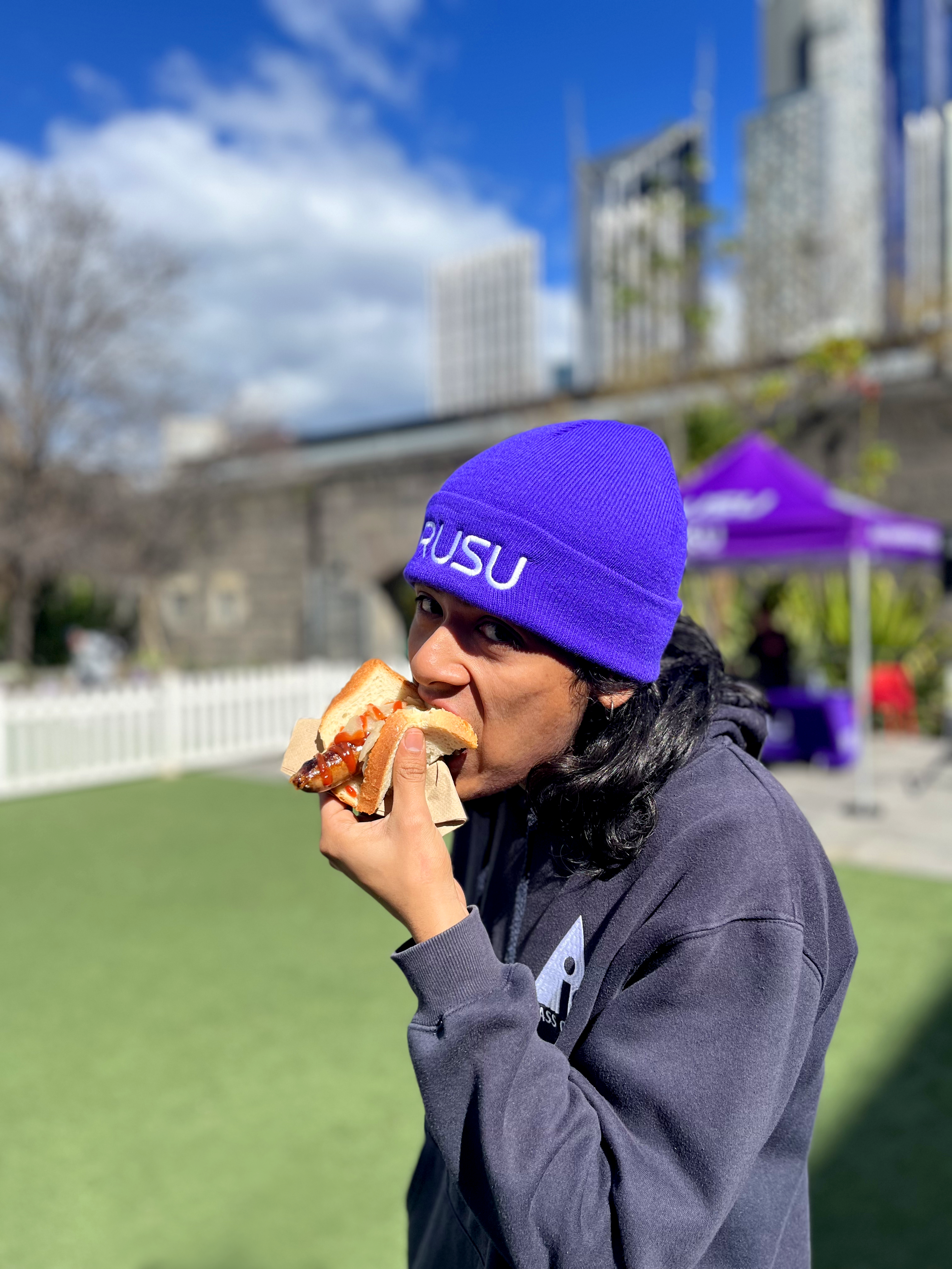 RUSU Beanie in purple with white RUSU embroidered logo modelled by male student outside on the green grass at RMIT University in the Melbourne CBD.