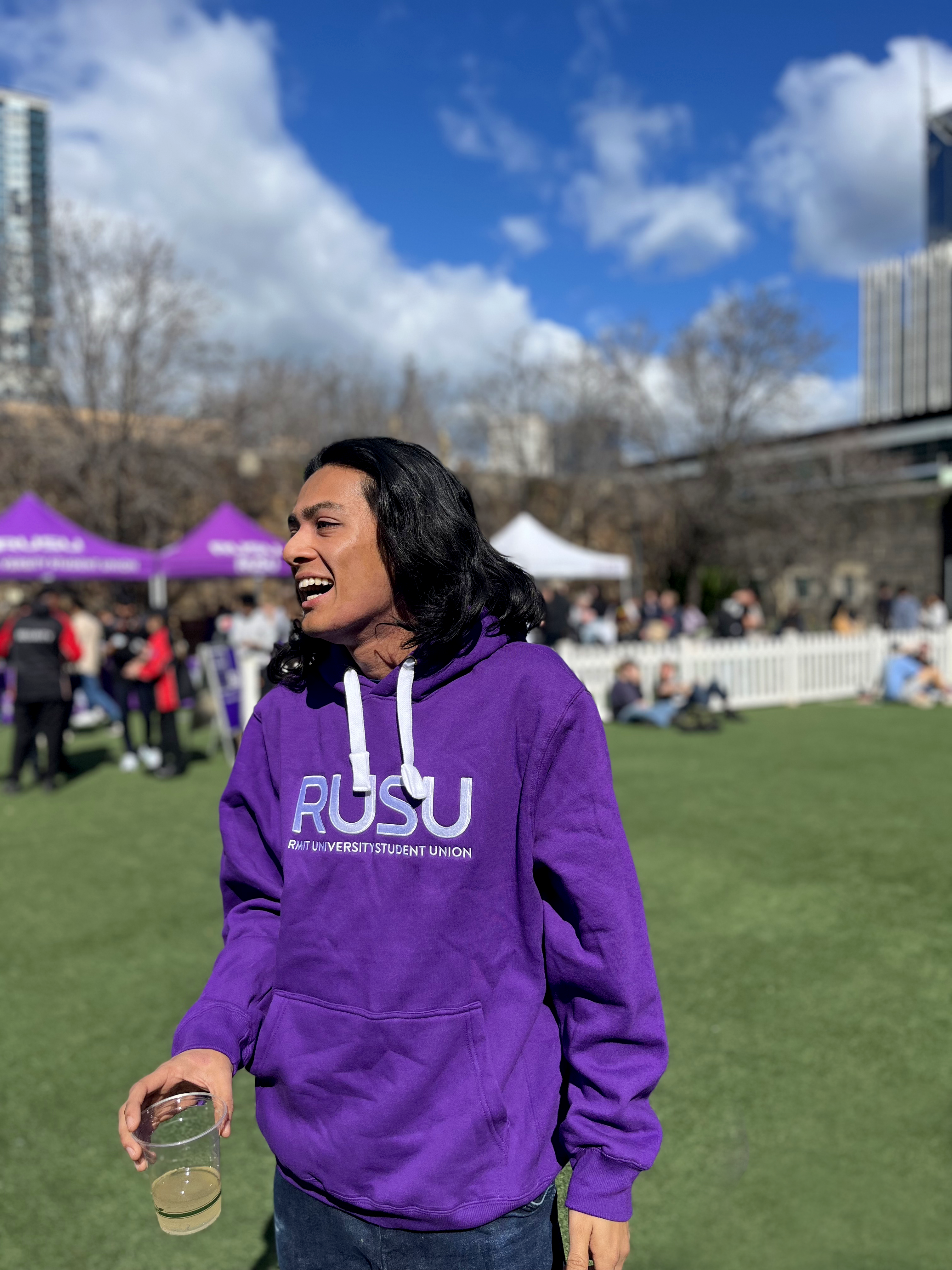 RUSU Hoodie in purple with large embroirdered white RUSU logo in centre chest. Modelled by student on Alumni Courtyard lawn at RMIT University in the Melbourne CBD.