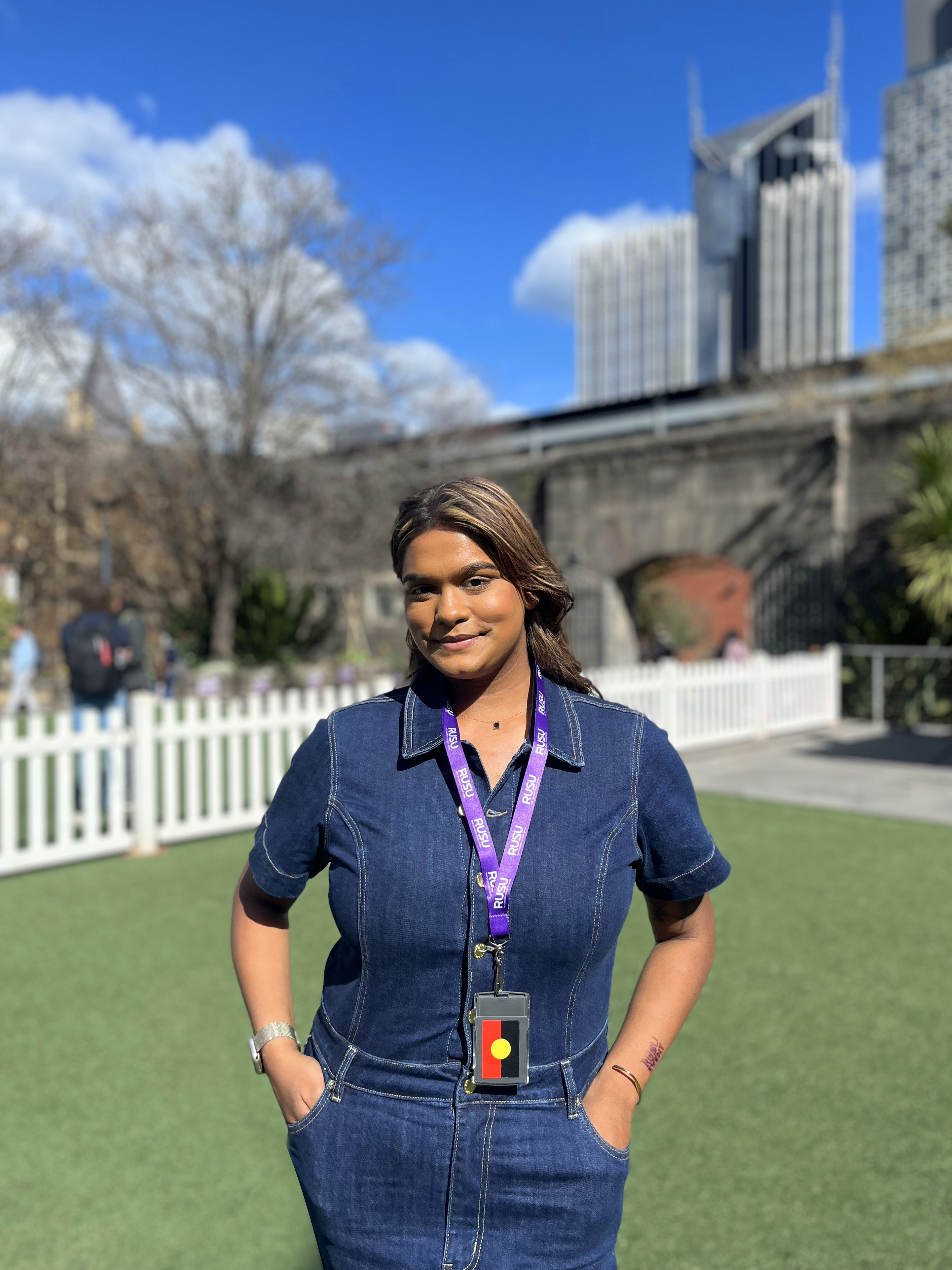 Model showcasing a purple RUSU lanyard from the RMIT Store, worn around the neck over a denim jumpsuit, in an outdoor setting with a cityscape. 