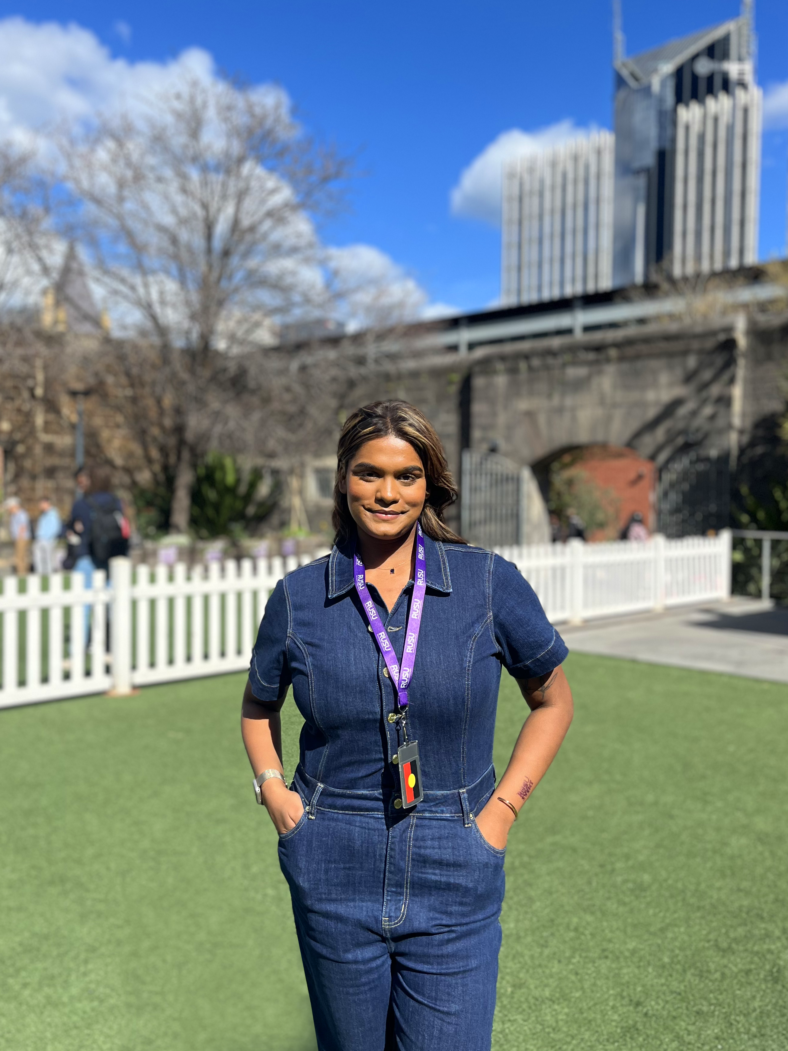Model in a denim jumpsuit wears a purple RUSU lanyard from the RMIT Store around her neck, standing outside against an urban backdrop.